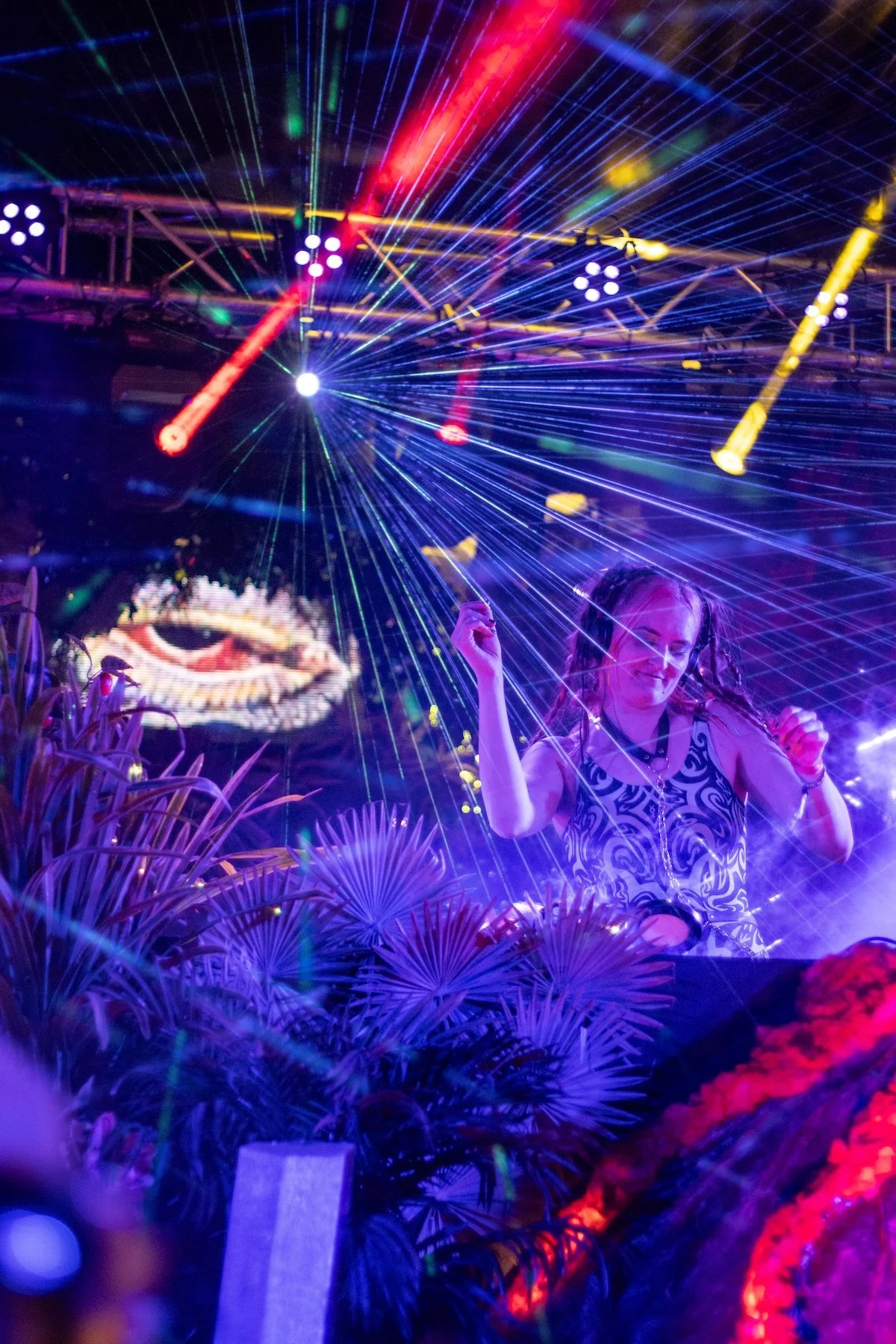 A woman dancing at a nightclub with colorful laser lights and digital visuals in the background, surrounded by plants.