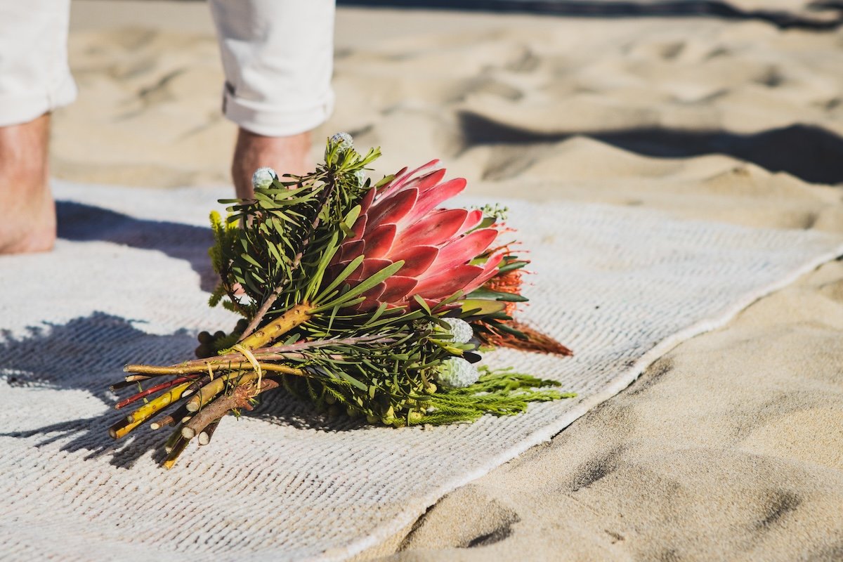 A wedding bouquet of pink protea flowers and greenery on a white cloth on the sand at the beach, with a person's legs visible in the background.