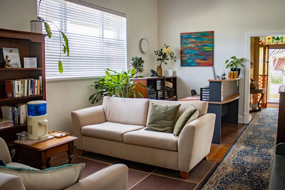 A cozy living room with a beige sofa, green pillows, and a beige armchair. A wooden coffee table with a water dispenser and glasses is beside the sofa. There are multiple plants, a bookshelf with books, and a small cabinet with decorative items. A co
