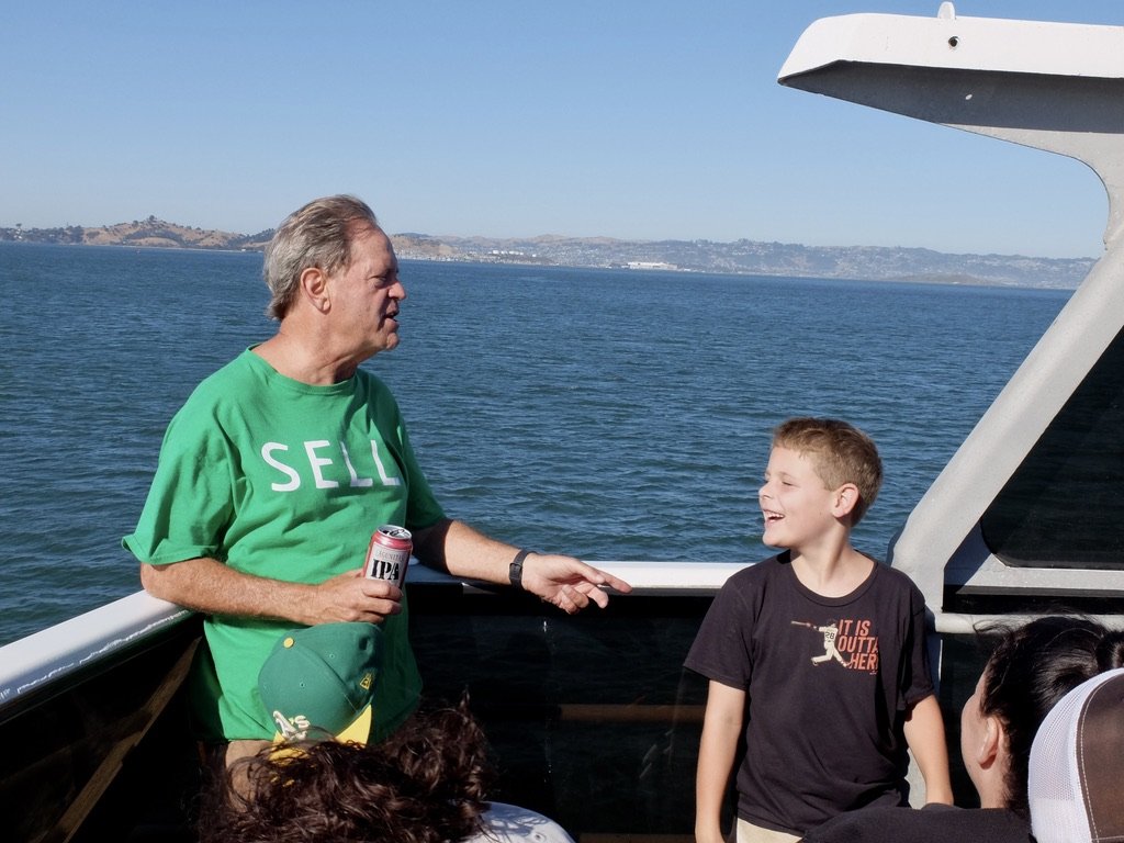  On the ferry to the San Francisco Oracle Giants Stadium to see a Giant’s vs. Oakland Athletics ball game.  Grandpa, an Oakland A’s fan, wanting the owner of the team to sell vs. moving the team to Las Vegas. Grandson a SF Giants fan. 