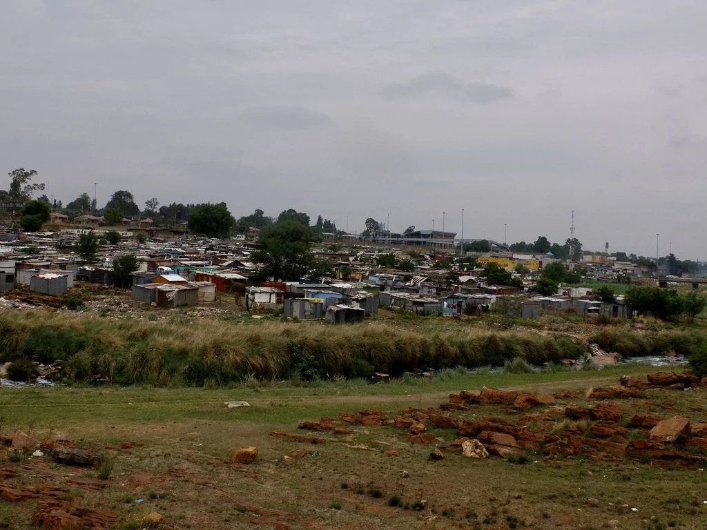  Gold mine tailings can be seen  in front of these shacks.  This area is temporary housing for immigrants.  At least there’s water &amp; electricity.  The government is desperately trying to house these people, many coming from Zimbabwe to find work 