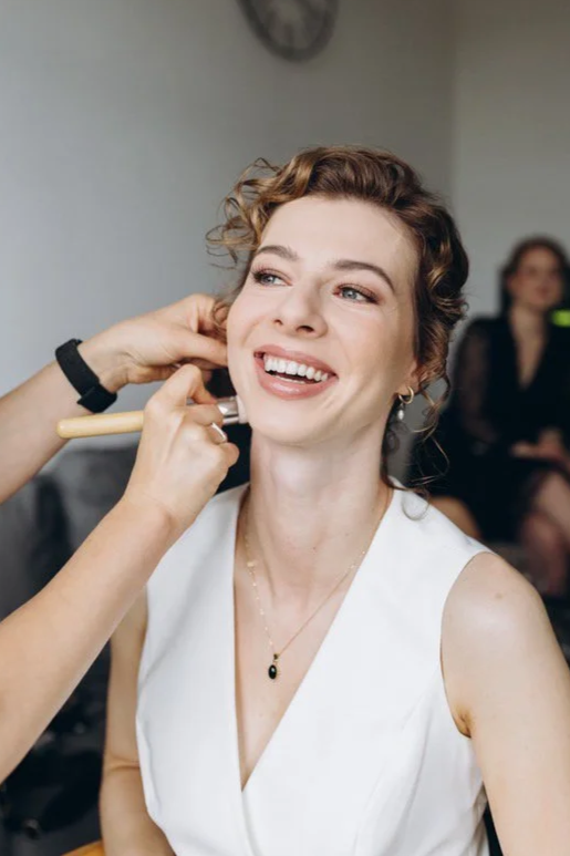 A woman with short curly hair wearing a white sleeveless top is having makeup or beauty beauty application done by a person using a brush, in an indoor setting. The woman is smiling and looking slightly to the right.