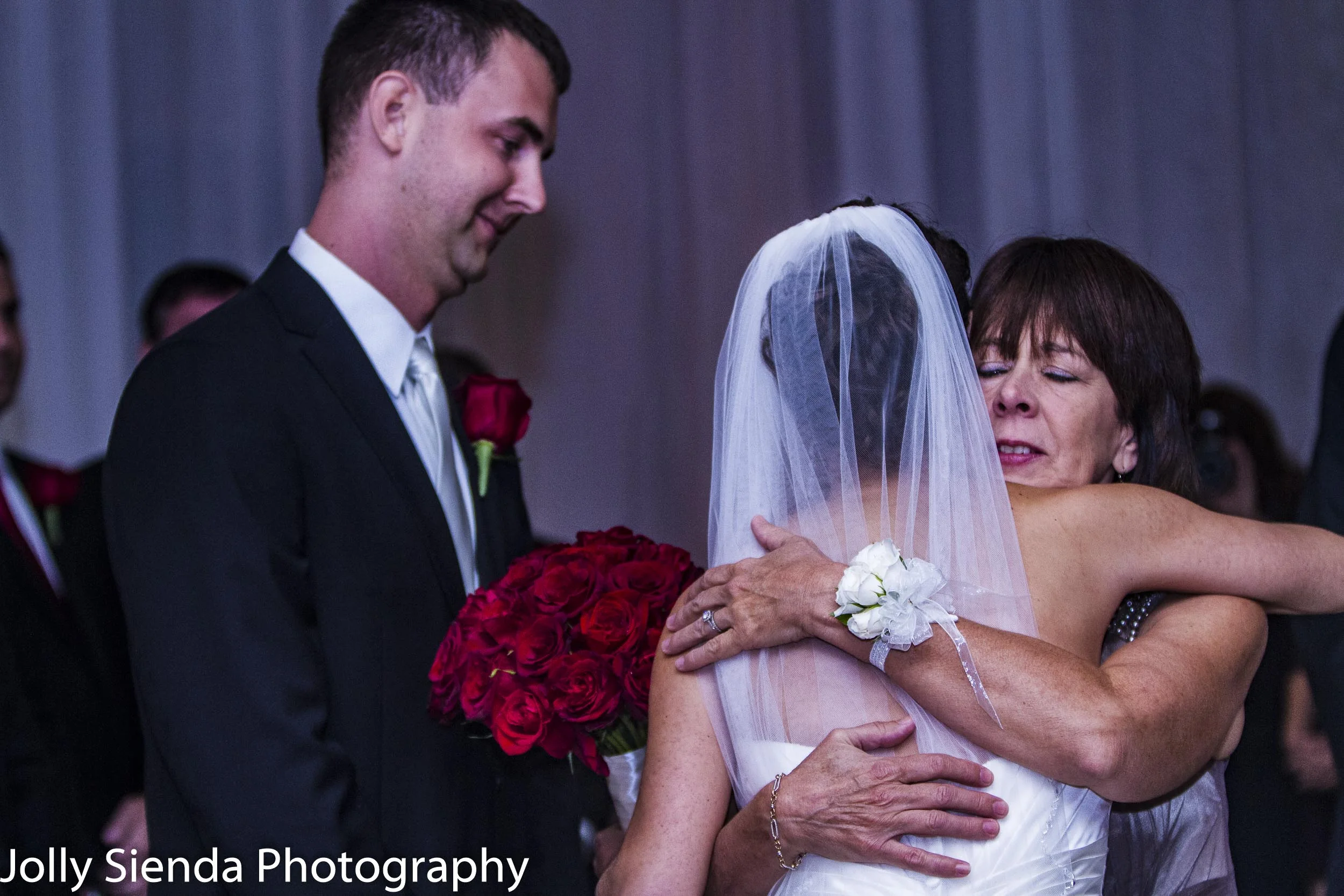 Bride at the alter gets a hug from her mother with the groom looking on and smiling
