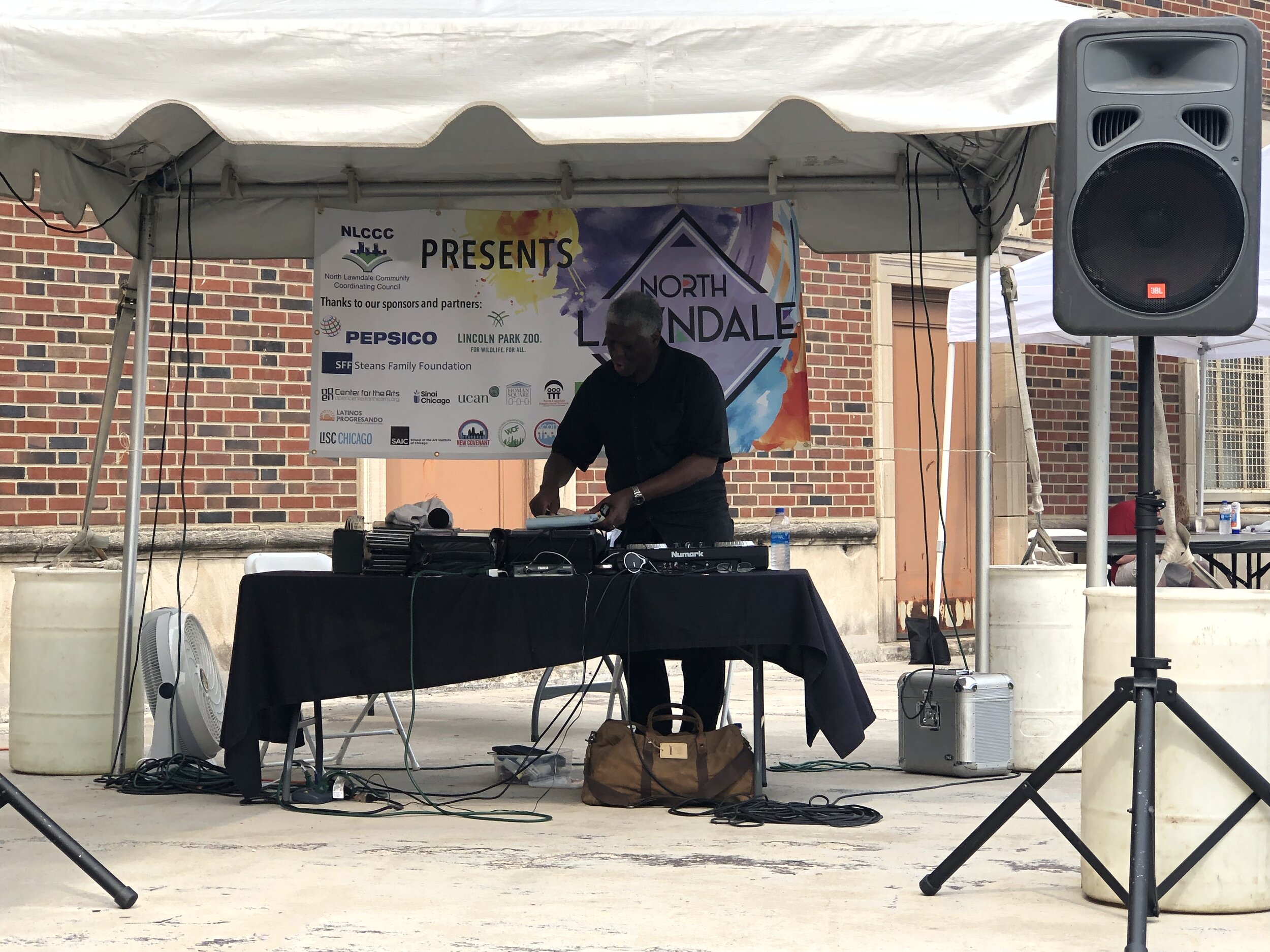 North Lawndale DJ playing music for people to dance and enjoy. In the back, there is a poster with sponsors of the North Lawndale arts festival.