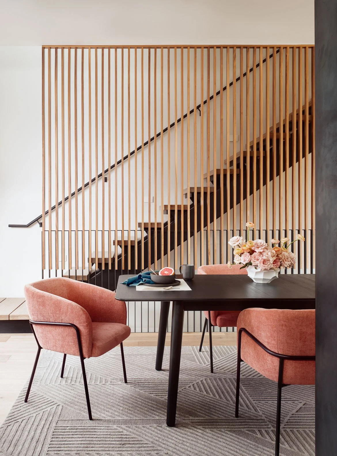 Modern dining room with coral chairs beside a wood slat staircase, Breathe Design Studio