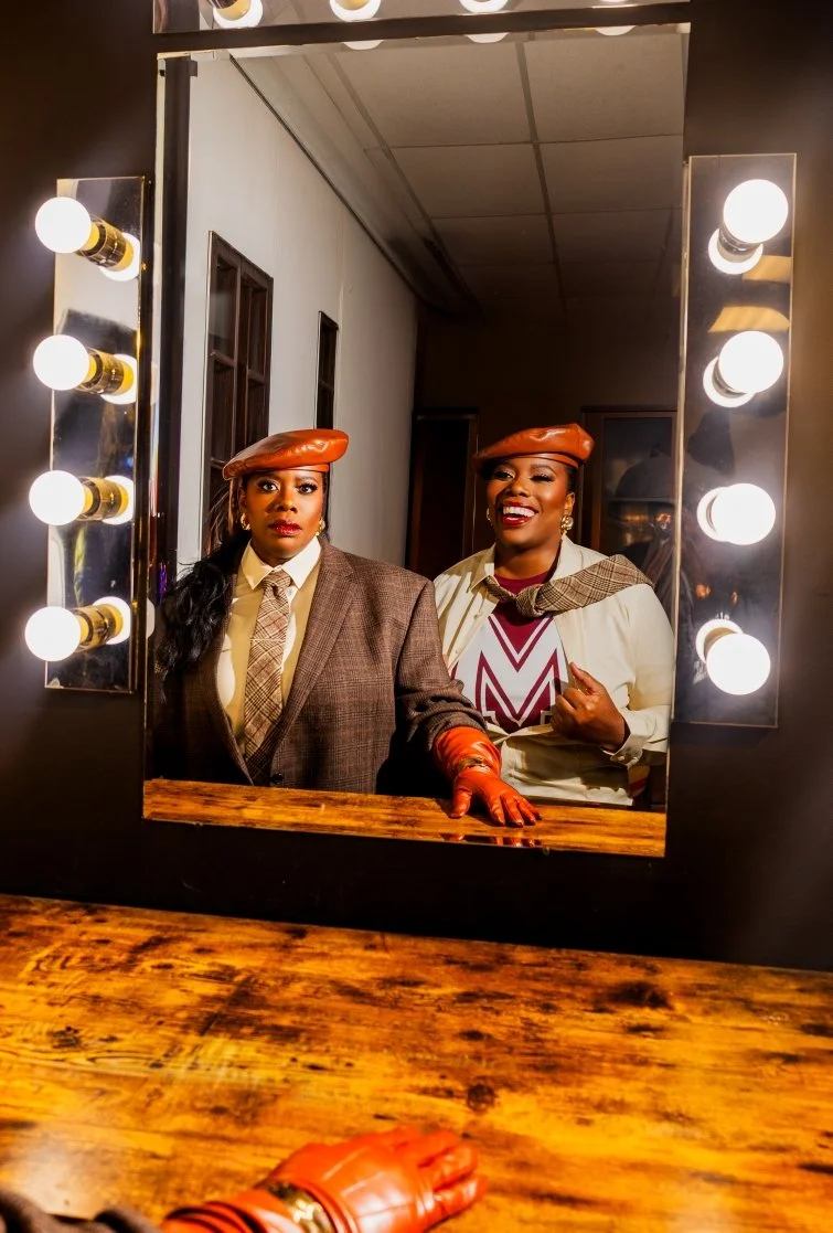 Two women dressed in vintage clothing and berets looking into a mirror with bright lights, smiling and posing for a photo.