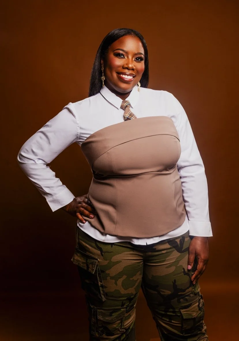 A woman with dark hair and dark skin smiling, dressed in a white shirt, beige corset-style top, plaid tie, camo pants, and gold earrings, standing against a brown background.