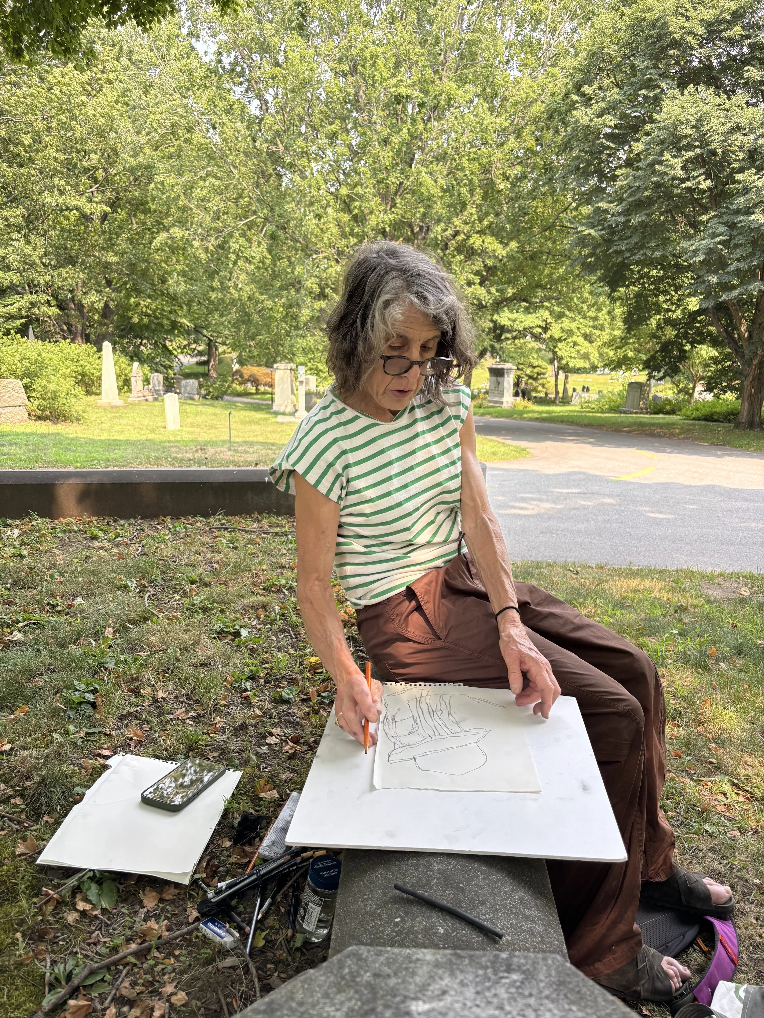 An elderly woman with gray hair, glasses, wearing a green and white striped shirt, is drawing on a large piece of paper outdoors in a park or graveyard area, seated at a white table with drawing tools, surrounded by trees and greenery.