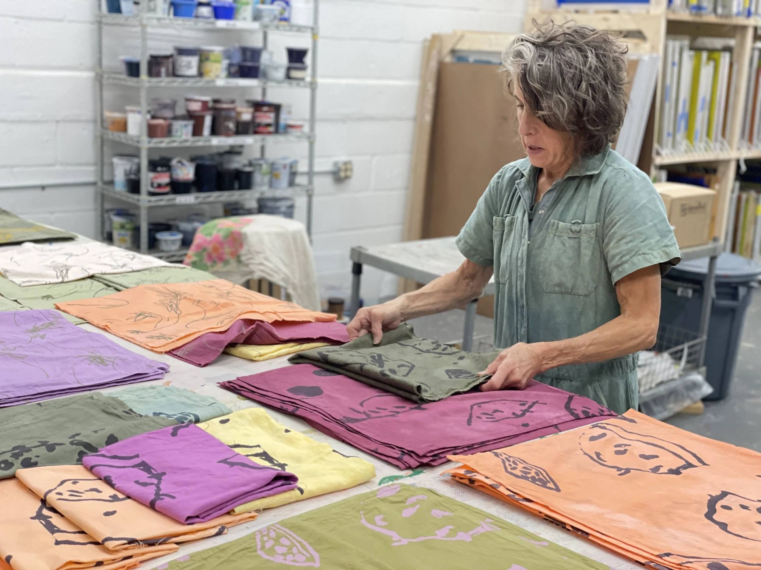 A woman with gray curly hair wearing a green shirt appears to be working with colorful fabric or paper designs on a table in an art studio or workshop. Shelves with various art supplies and materials are visible in the background.