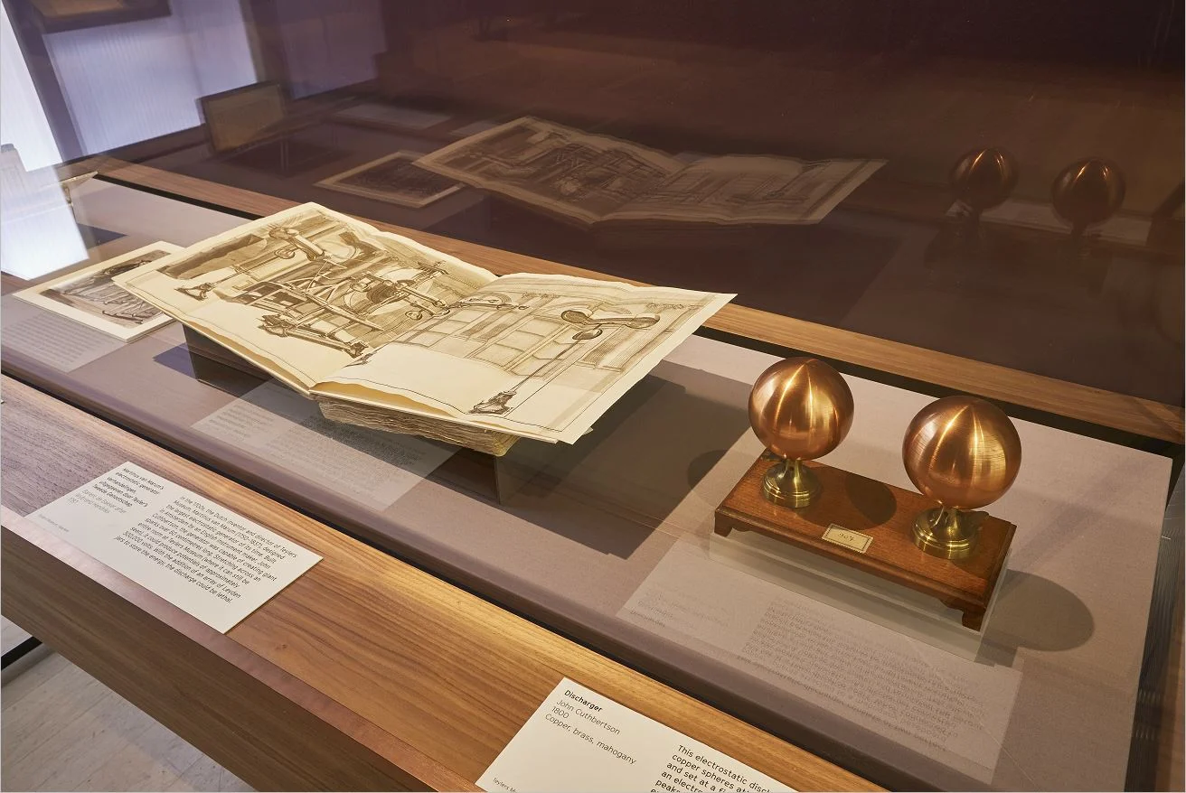 An exhibition display case containing an open book with an illustration of 19th century electrical apparatus. Next to the open book is an electrostatic discharger.