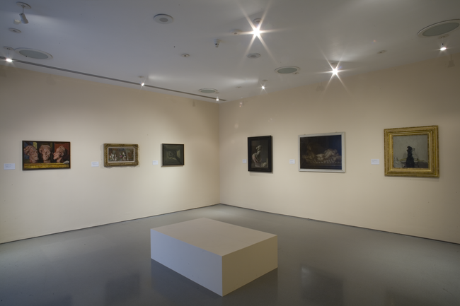 An exhibition of paintings on two walls of a gallery. A square bench is in the middle of the room.