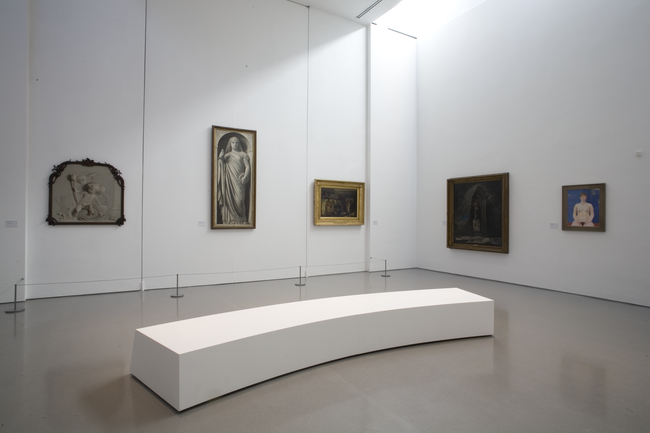 Exhibition space with two walls displaying paintings. There is a large and curvy rectangular  bench in the middle of the room.