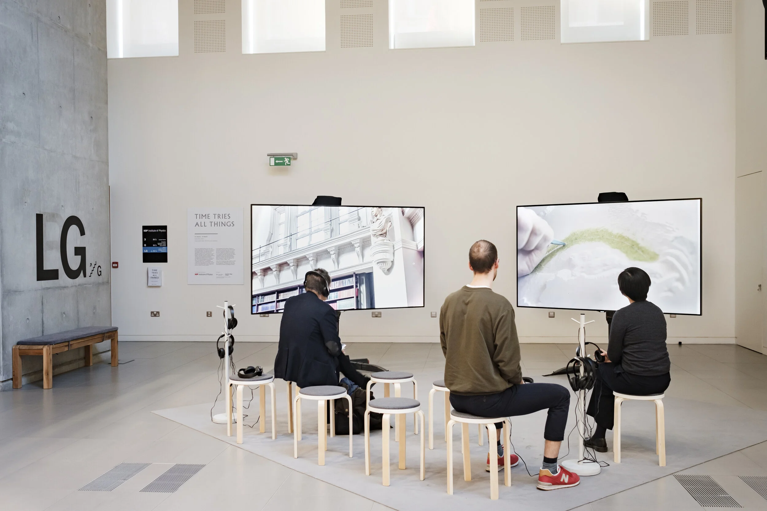 A two-screen film installation inside a gallery being watched by three seated people
