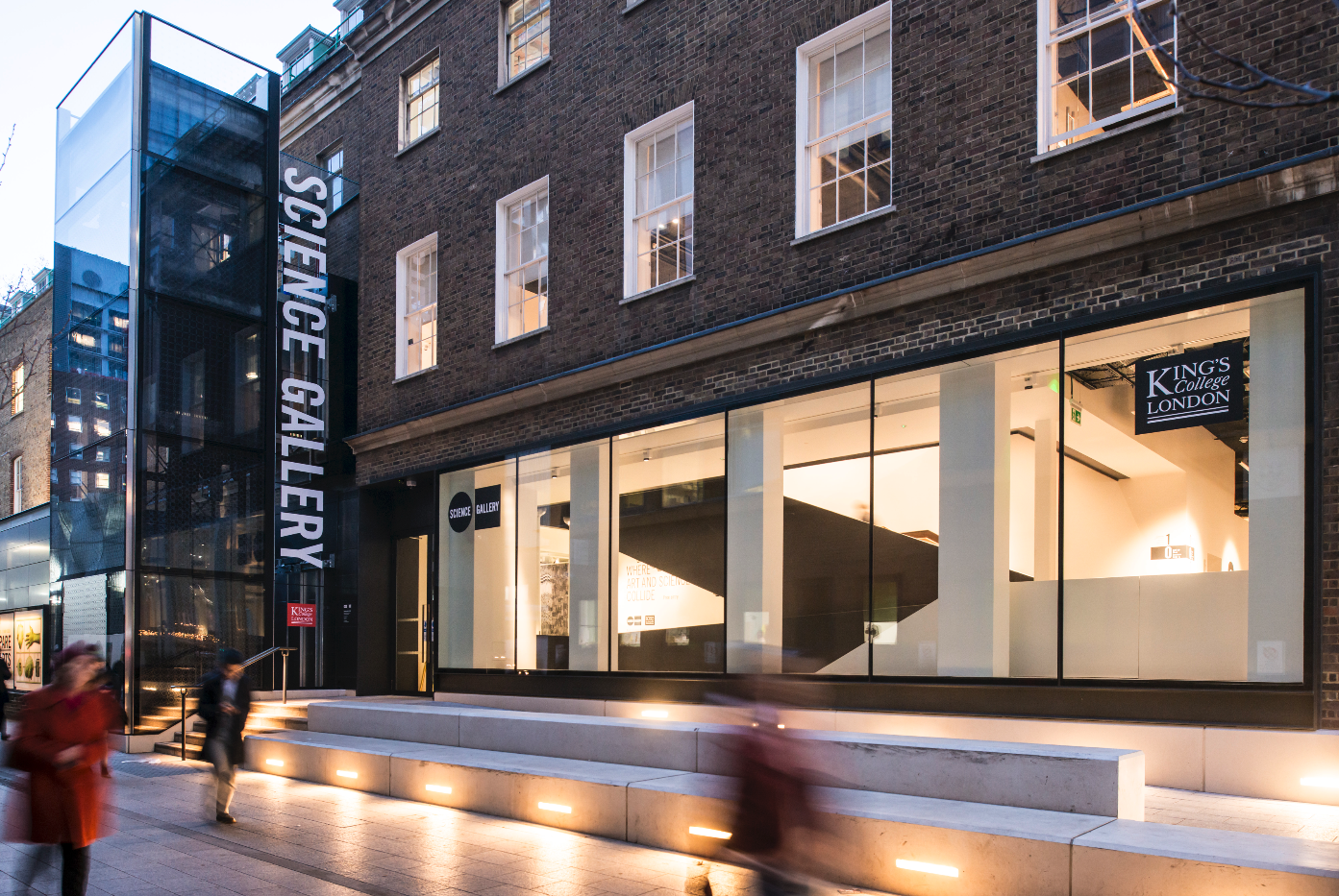The facade of Science Gallery London, a three storey building with a glass facade on the ground floor. A broad set of steps are lit up outside.