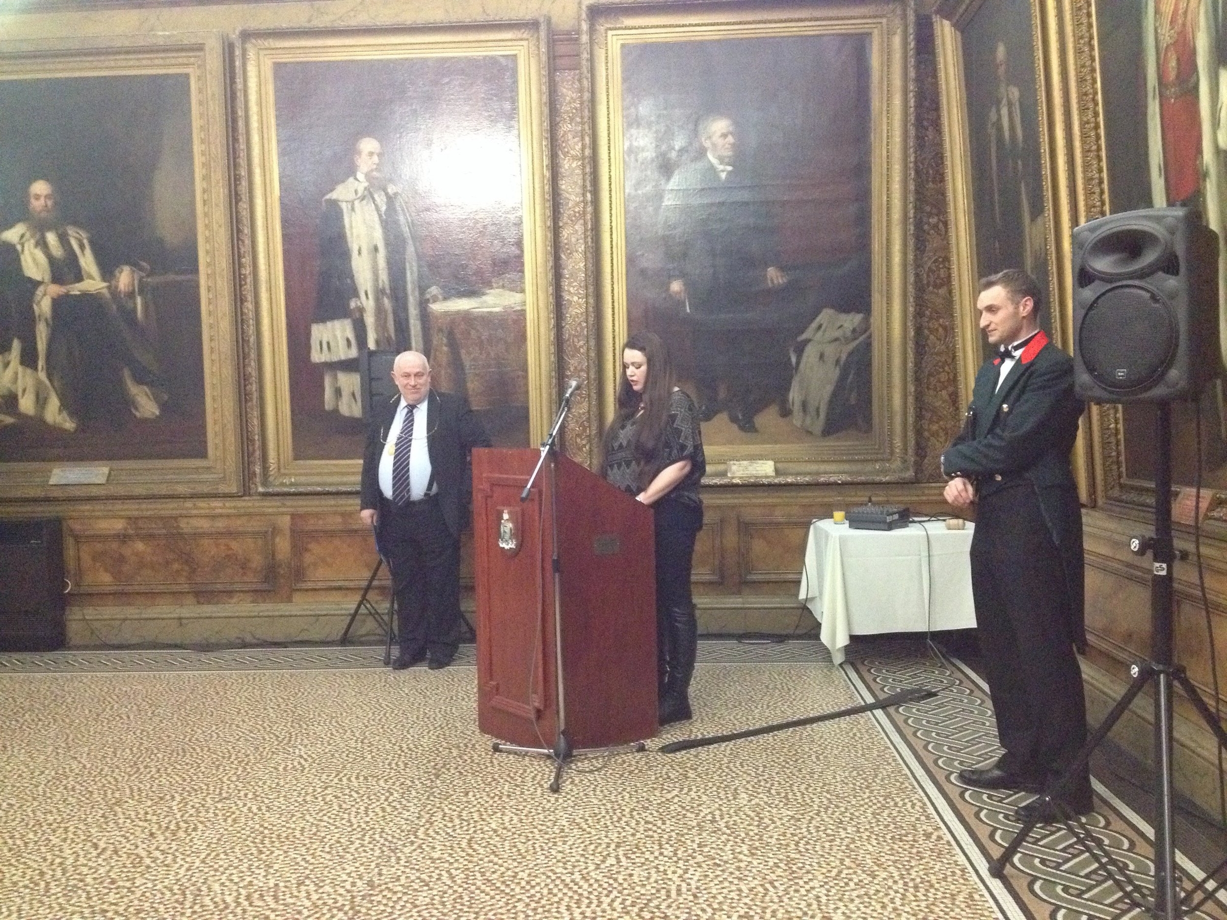 Speaker at a podium in the city Chamber with several large portraits behind her. There are two well dressed men at either side.