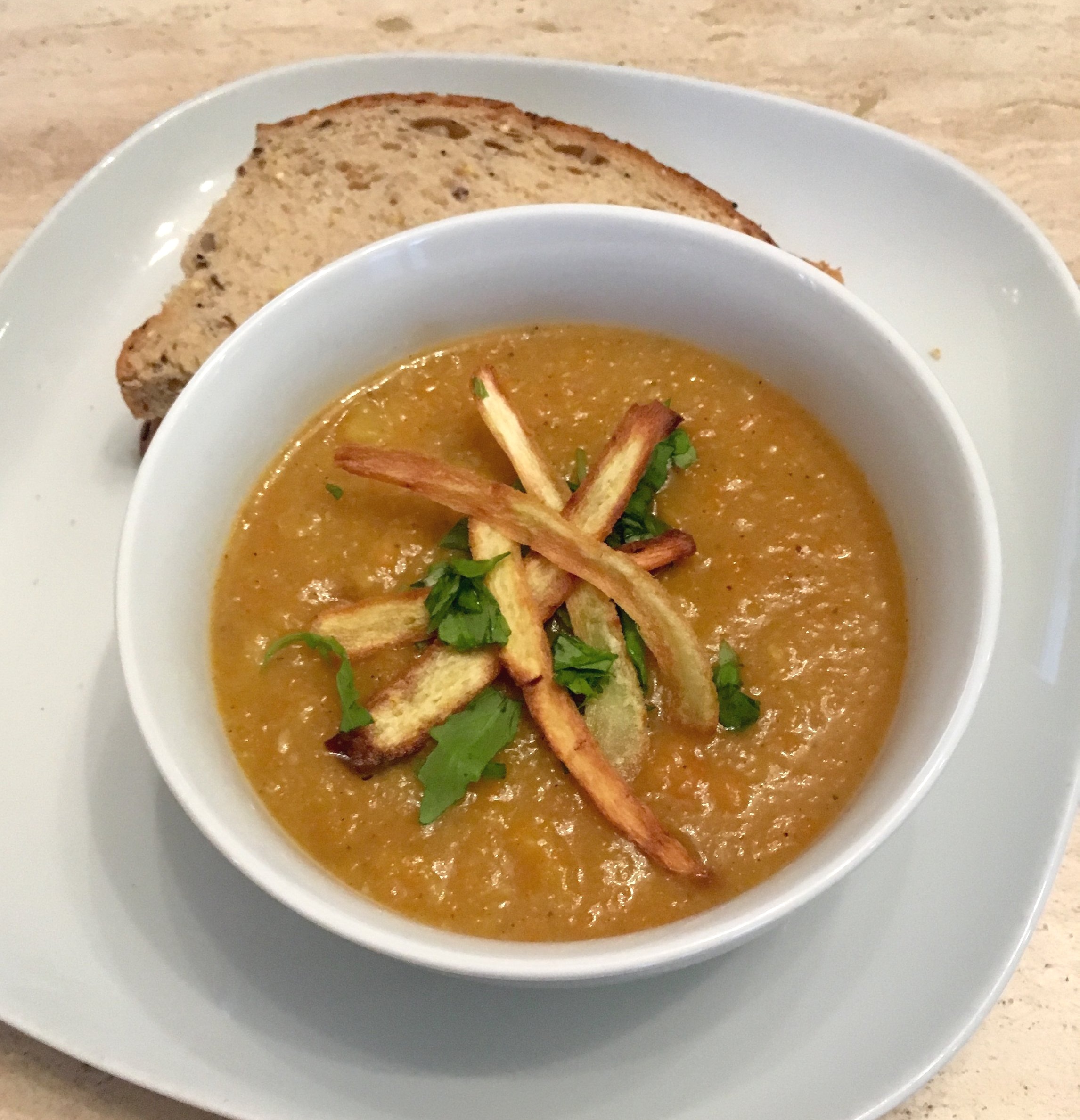 A bowl of carrot and parnsip soup with crispy parsnips and spinach on top. the Bowl is placed on top of a larger plate with 2 triangles of brown seeded bread.