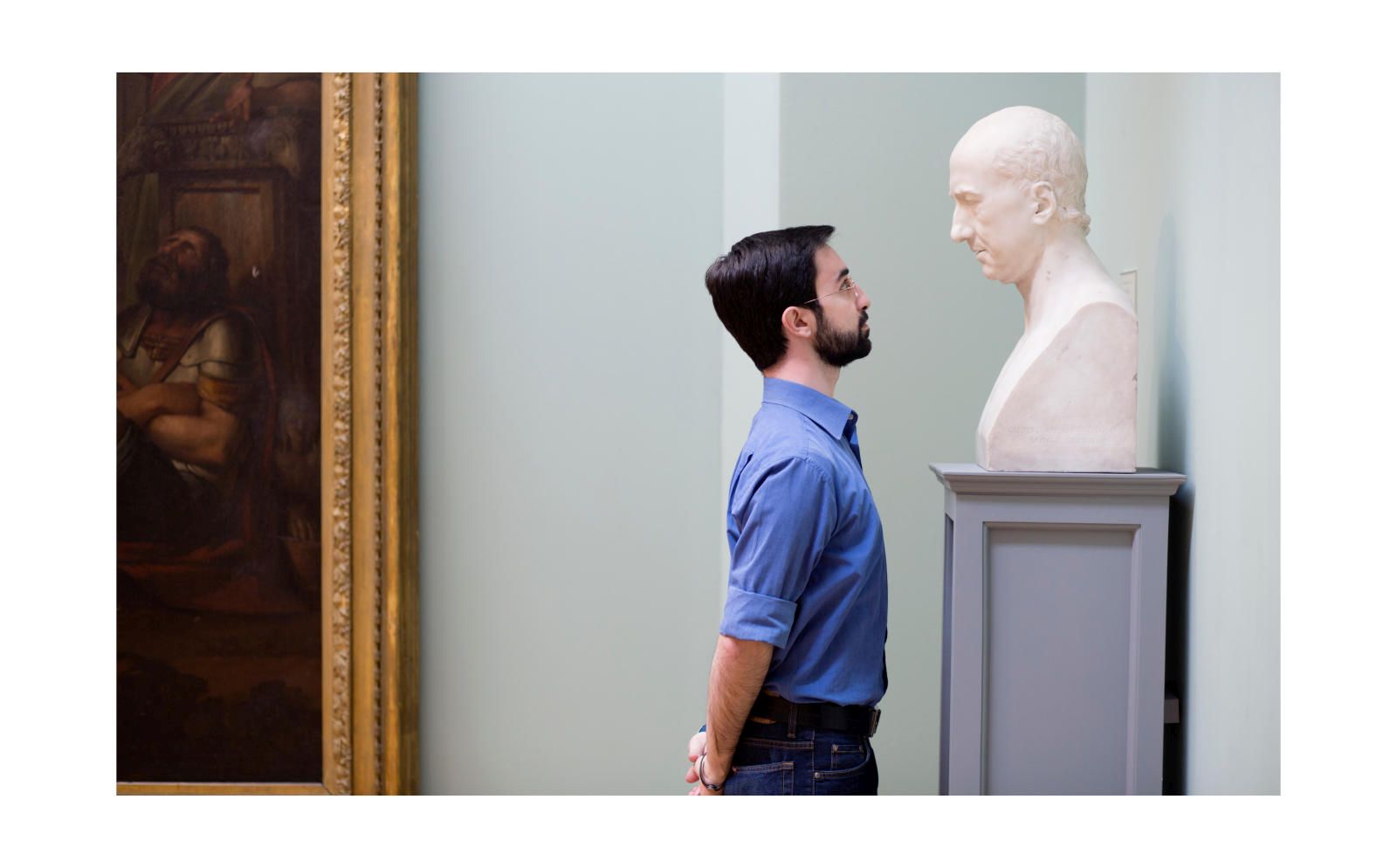 Man in a blue shirt staring eye to eye with a marble bust.