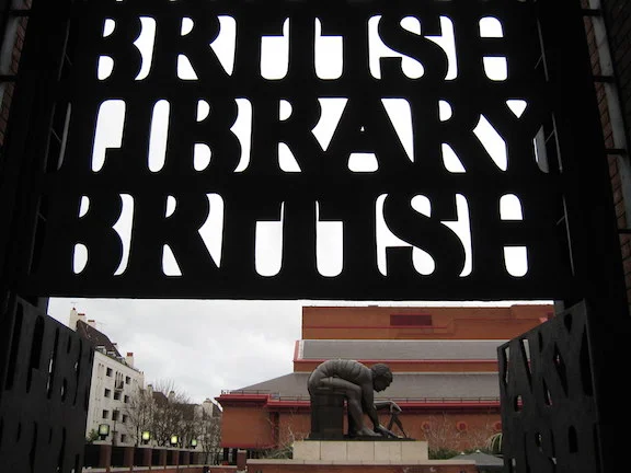 [Image Description: A sign that reads ‘British Library/British’ is shown in front of a statue of a man using a protractor. These are outside the British Library.]