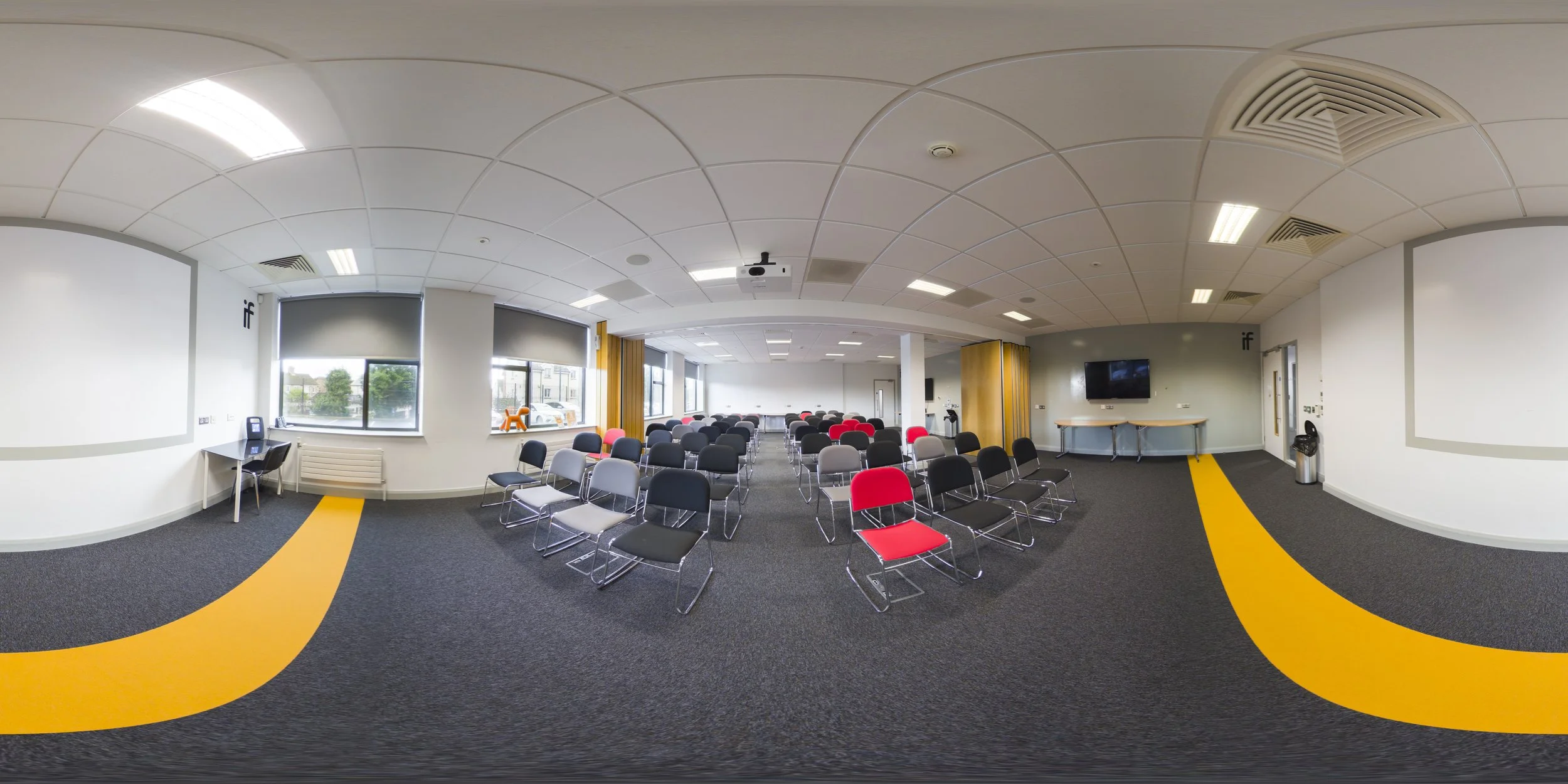 A spacious conference room with rows of chairs, some red and black, facing a large screen at the front. Large windows on one side let in natural light, and the ceiling has a grid pattern with installed lighting. There's a TV on the wall and a couple of tables at the sides, with a black mailbox or speaker on a table near the window.
