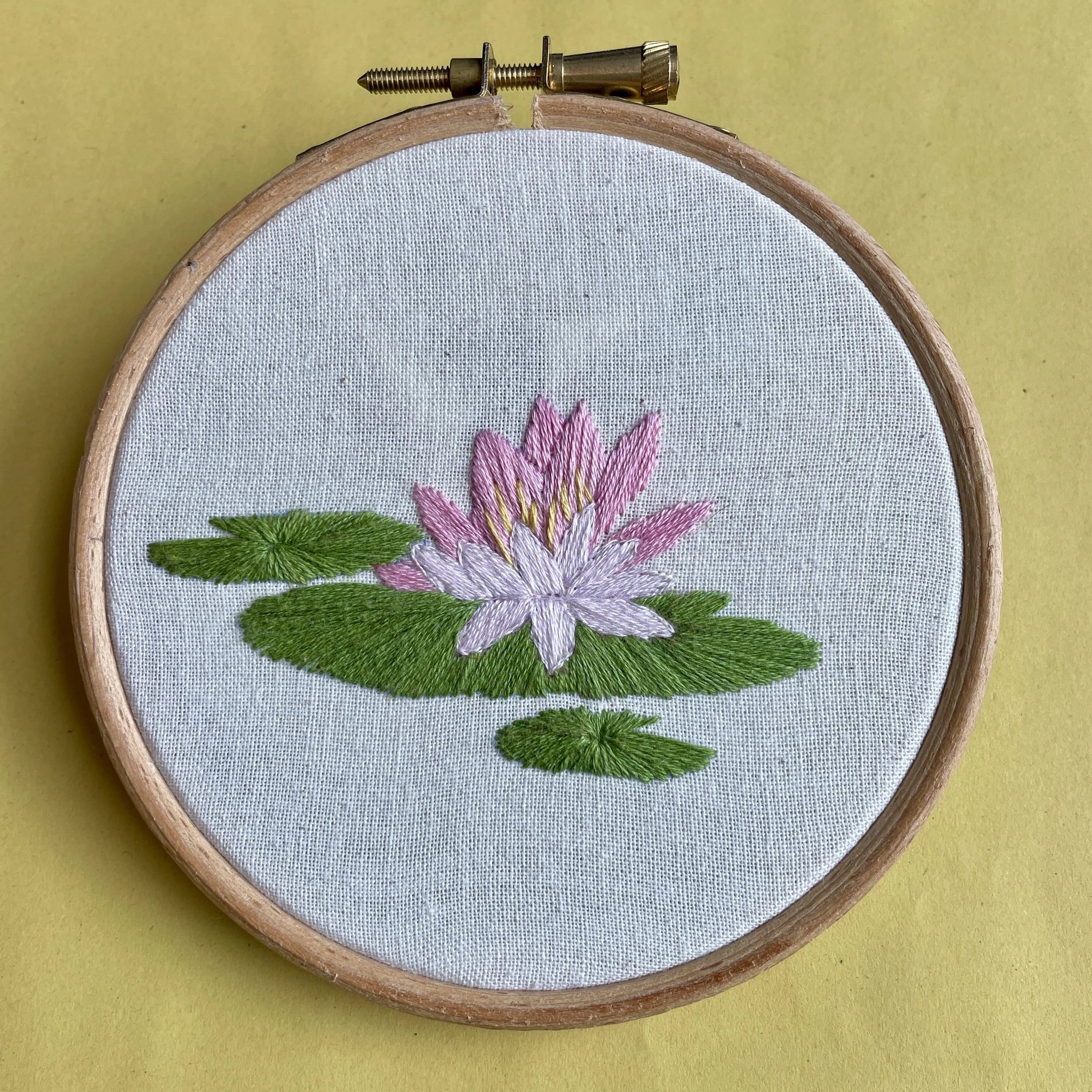 A picture of an embroidery hoop. In the hoop is calico fabric and on the fabric is stiched a pink waterlily design