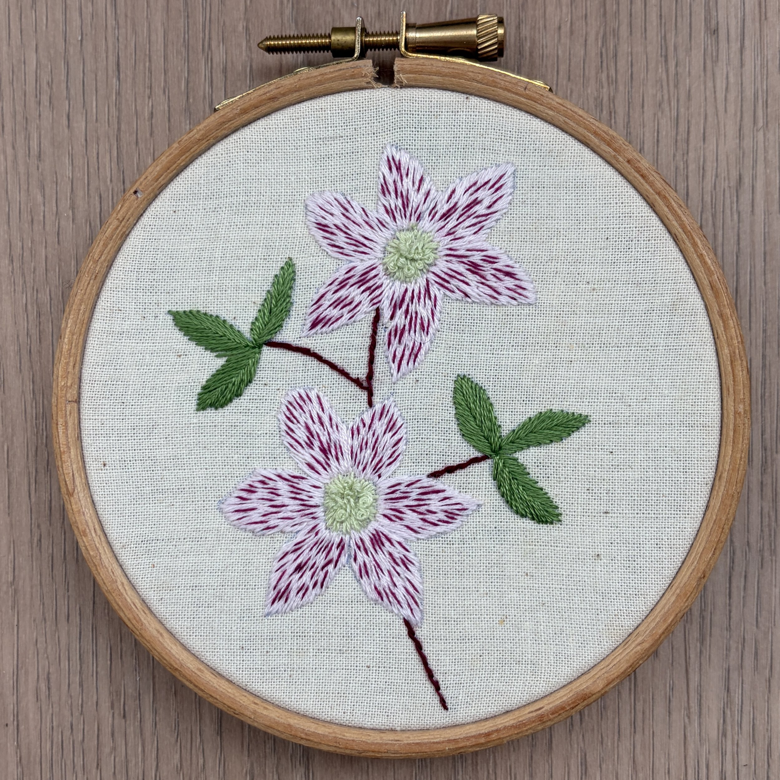 A picture of an embroidery hoop on a wooden background. In the embroidery hoop is some calico fabric and on the fabric is stitched a pink clematis with two flowers and two leaves.