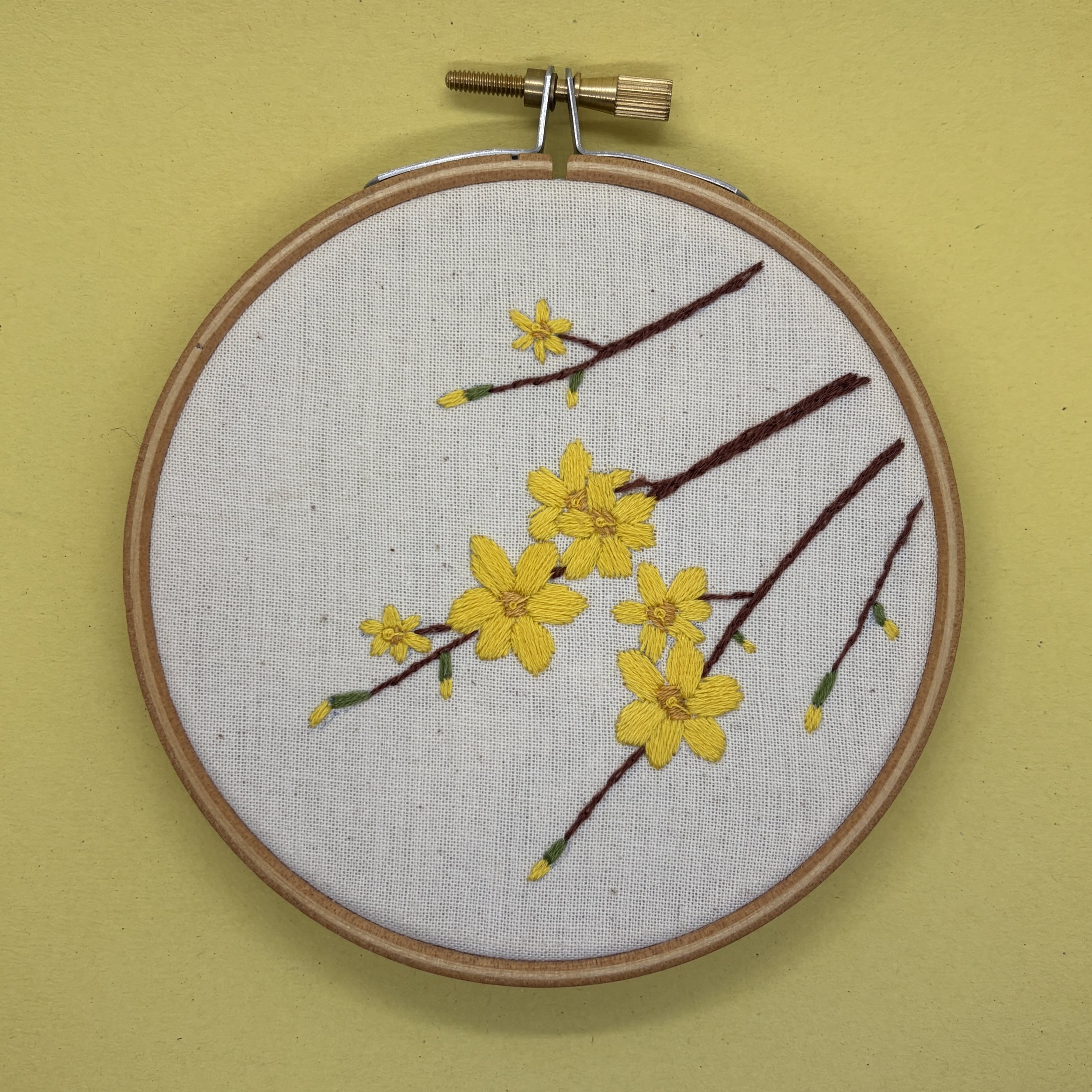 On a yellow background there is a wood embroidery hoop. In the hoop is calico fabric and on this is stitched four twigs, each twig has some yellow winter jasmine flowers stitched on it