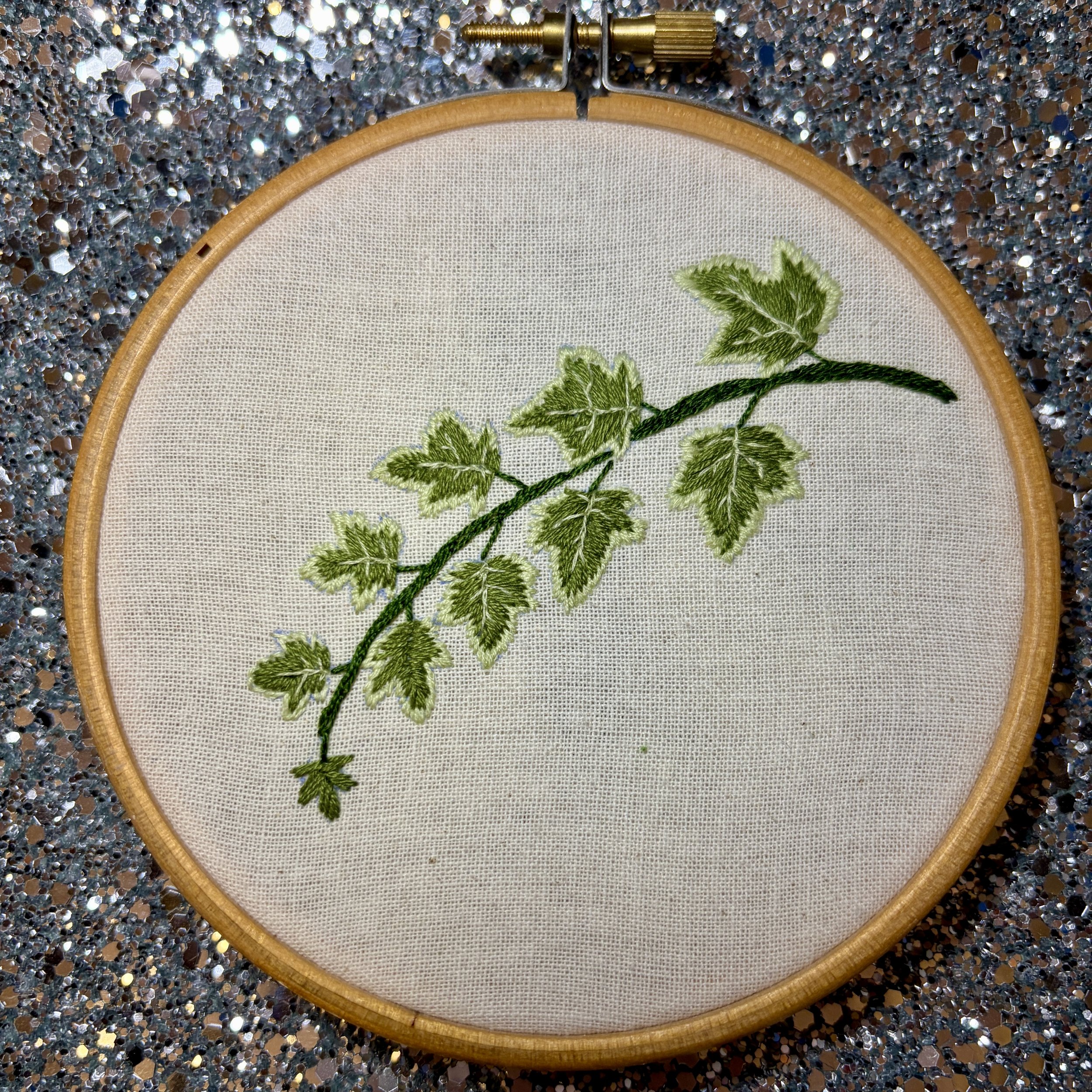A picture of an embroidery hoop on a shiny silver background. In the embroidery hoop is calico fabric and on the fabric is stitched a sprig of ivy