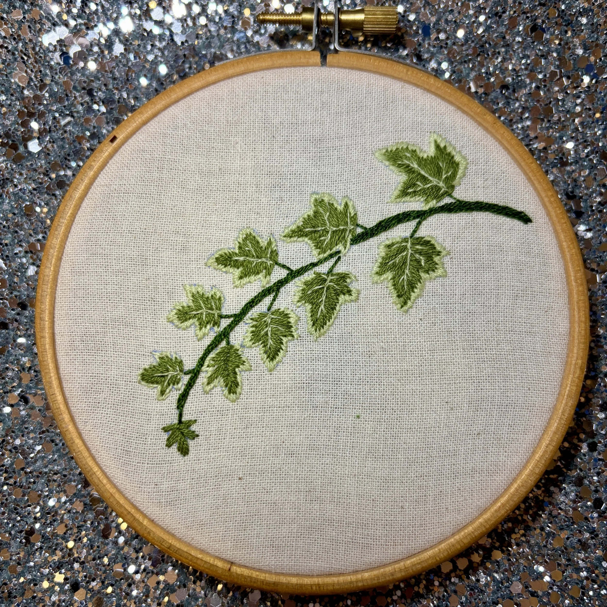 A picture of an embroidery hoop. In the hoop is calico fabric and on the fabric is stiched a sprig of ivy