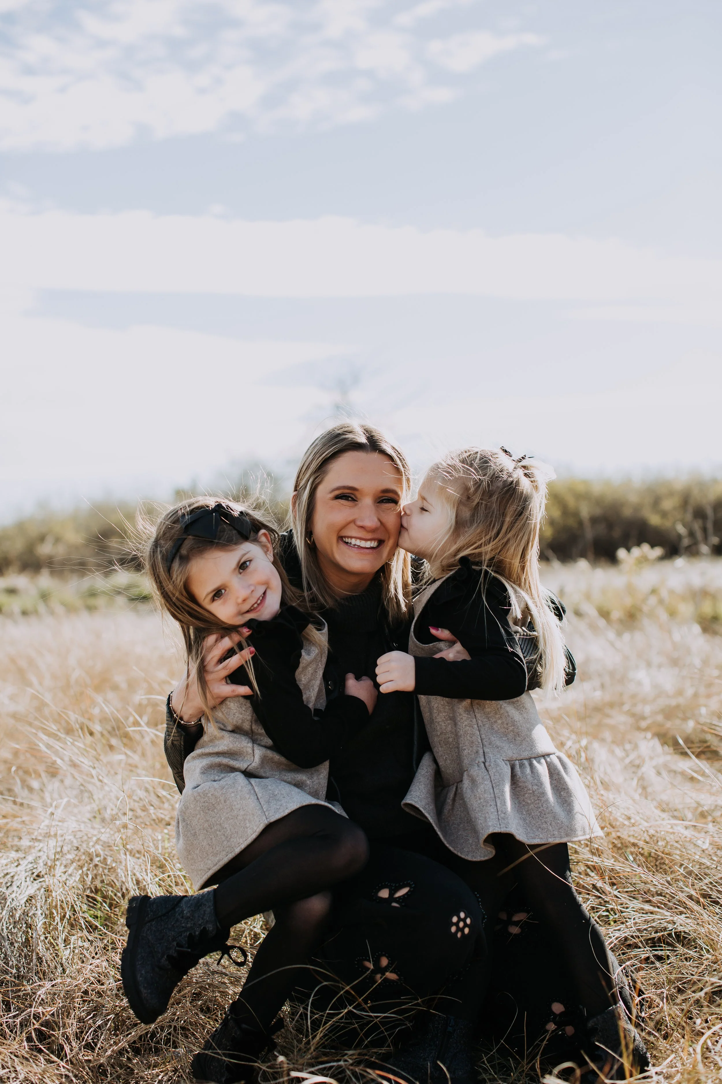 A woman with two young girls in a grassy outdoor setting, with the girl on the right giving the woman a kiss on the cheek and the girl on the left smiling while hugging her. All are dressed in black and grey clothing, with the background showing a pa