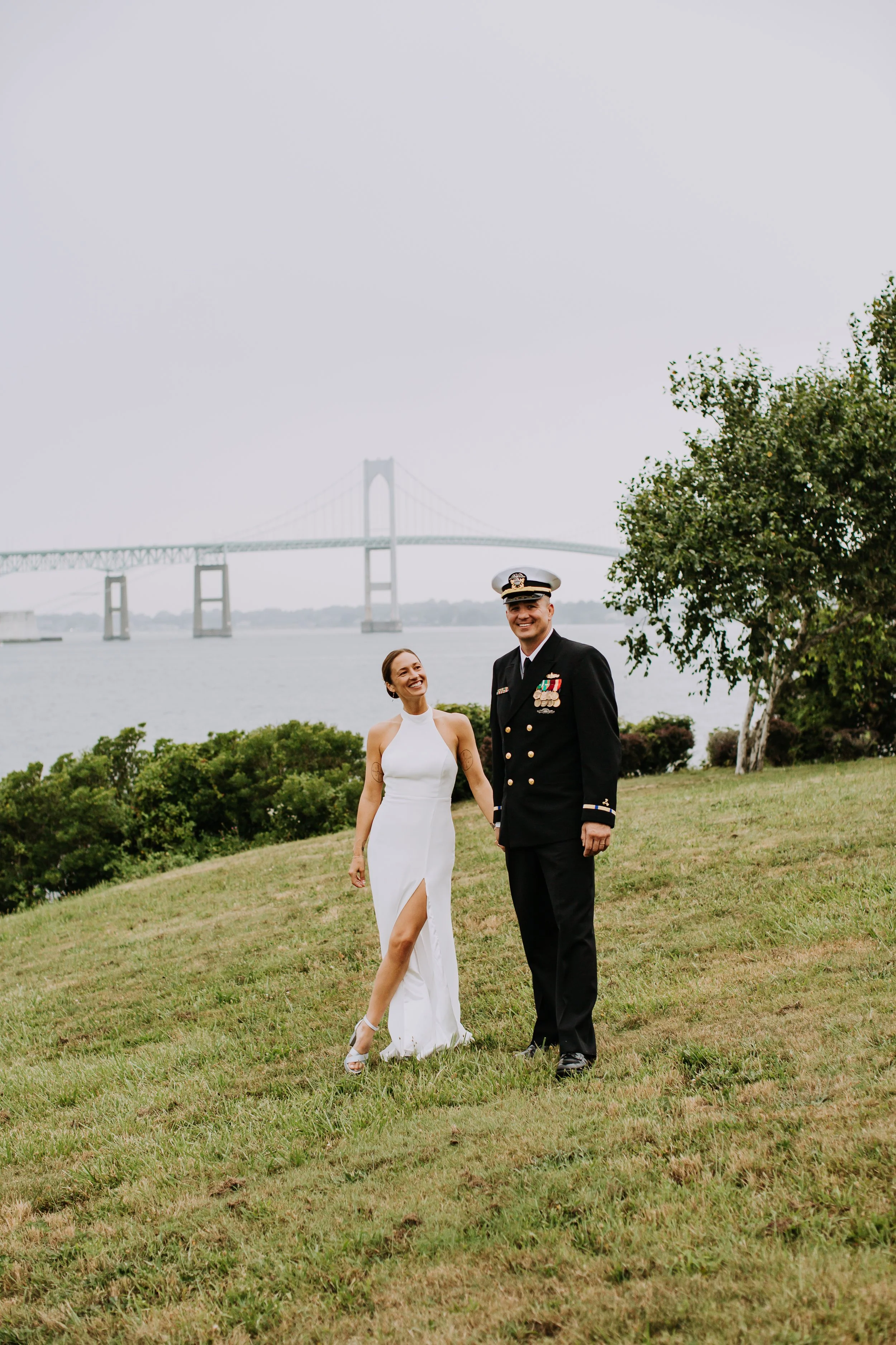 A couple in wedding attire, holding hands and smiling, standing on grass with a bridge and water in the background.