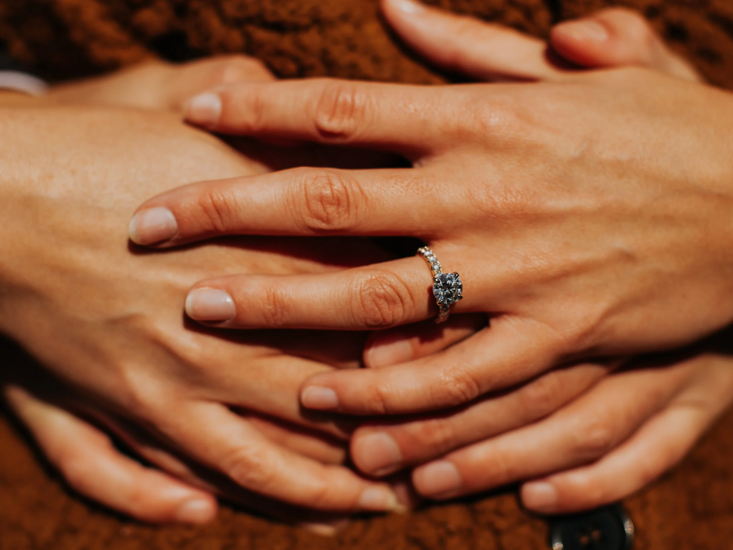 Close-up of two hands with an engagement ring featuring a large diamond on the ring finger, resting on a textured brown surface.