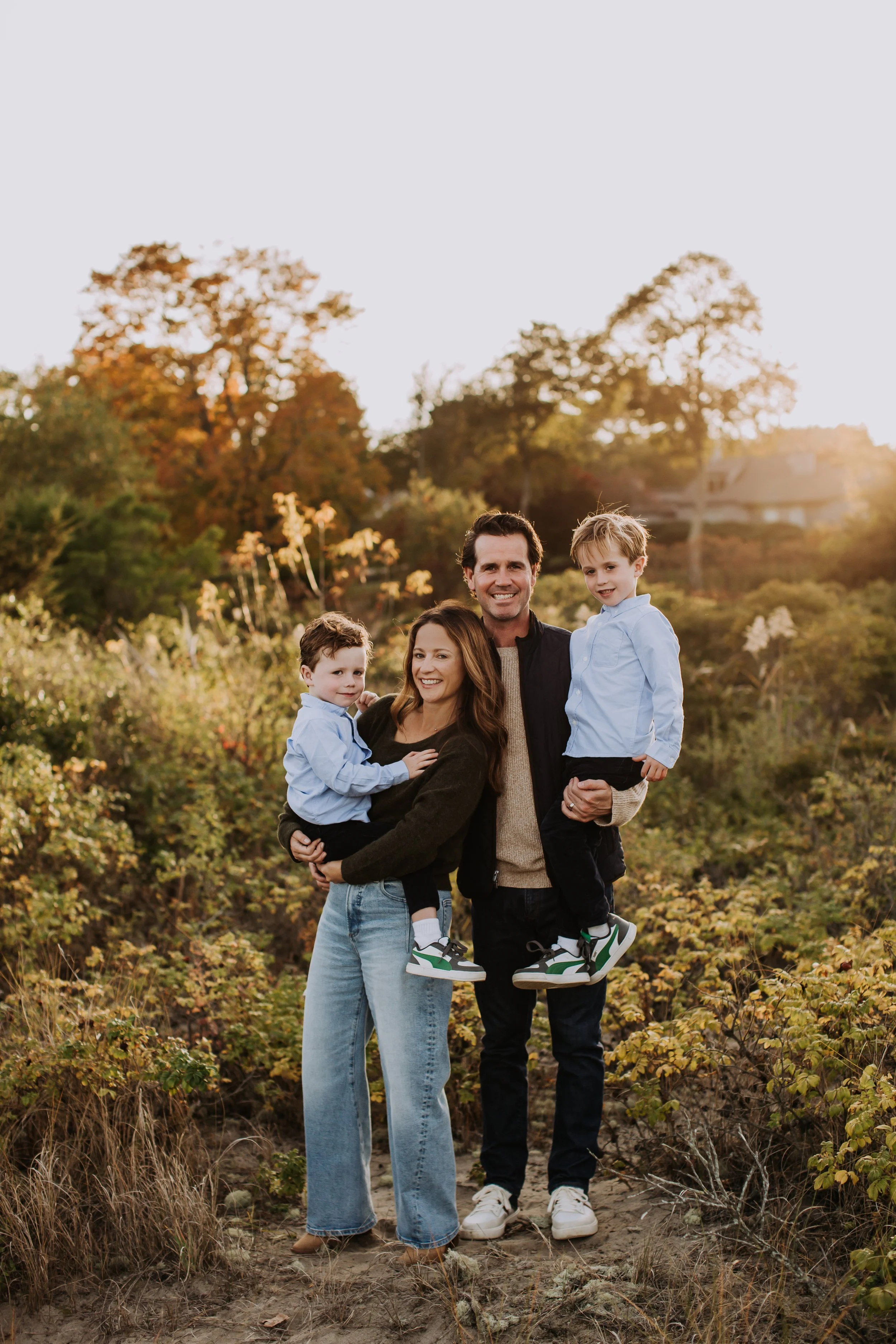 A family of four outdoors during sunset, standing on a dirt path surrounded by autumn foliage, smiling at the camera.