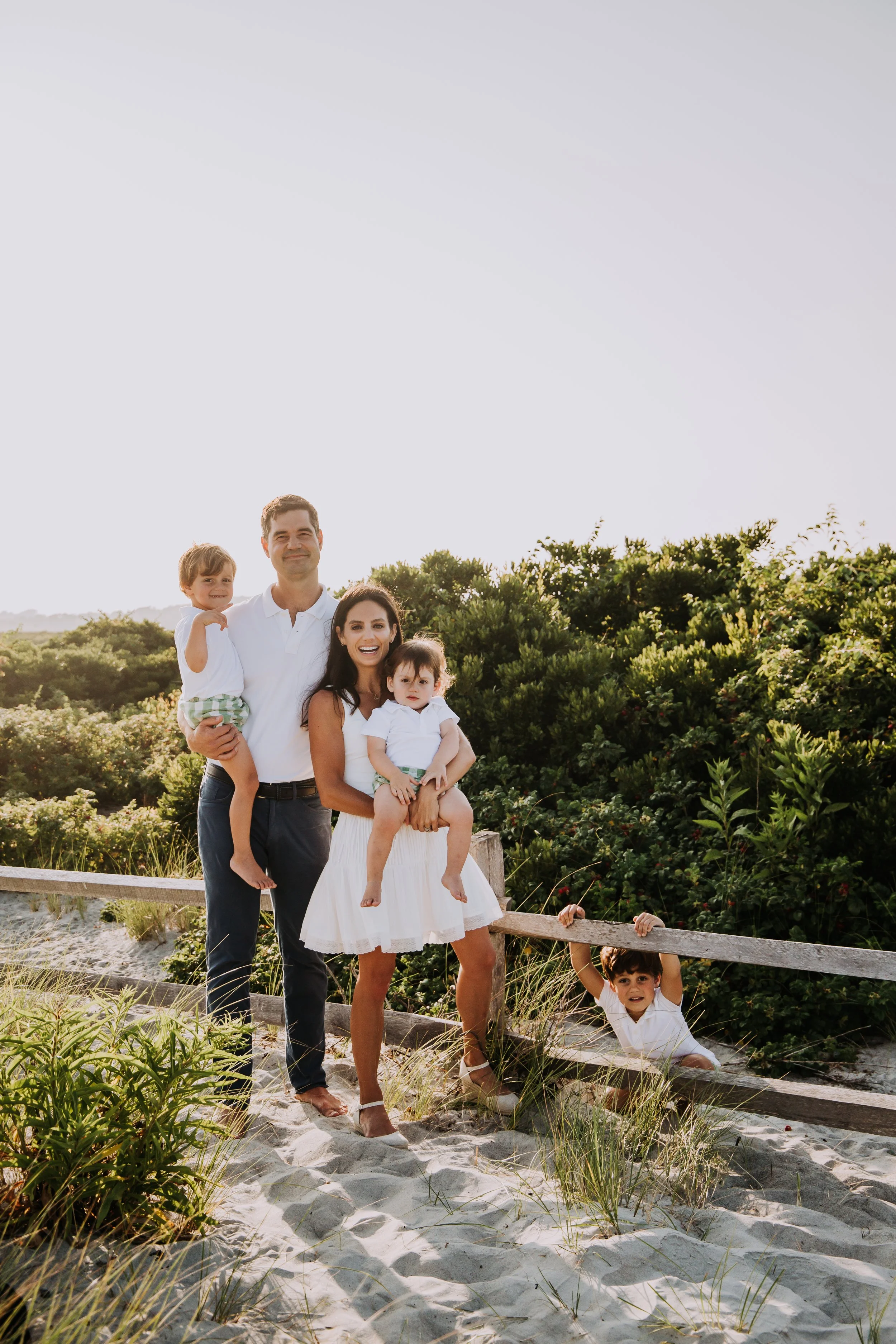 Family of five standing on sandy beach near green bushes and railing, smiling at camera during daytime.