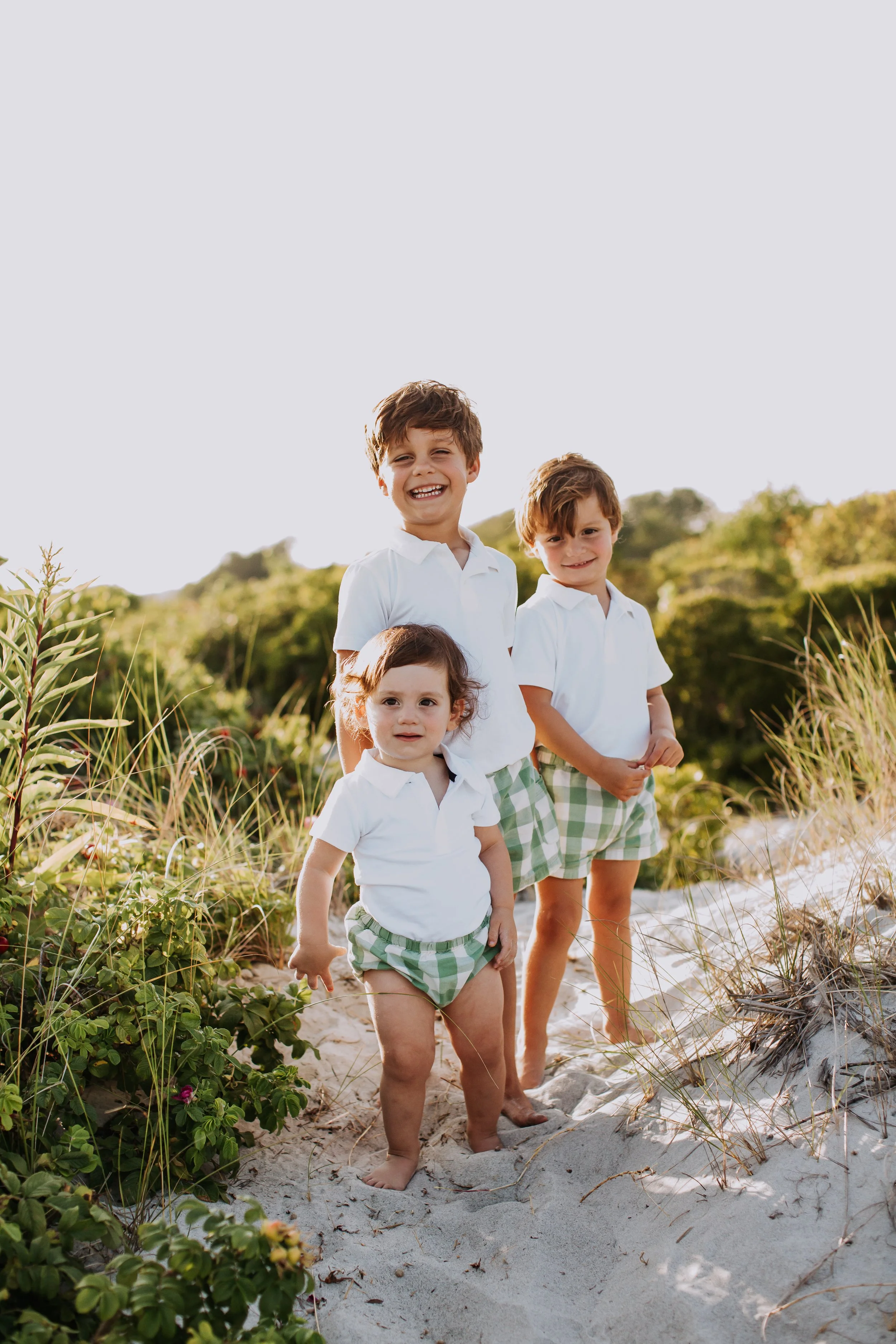Three young children, two boys and a girl, standing barefoot on a sandy beach with greenery and dunes in the background, all wearing white shirts and green checkered shorts, smiling and enjoying a sunny day.