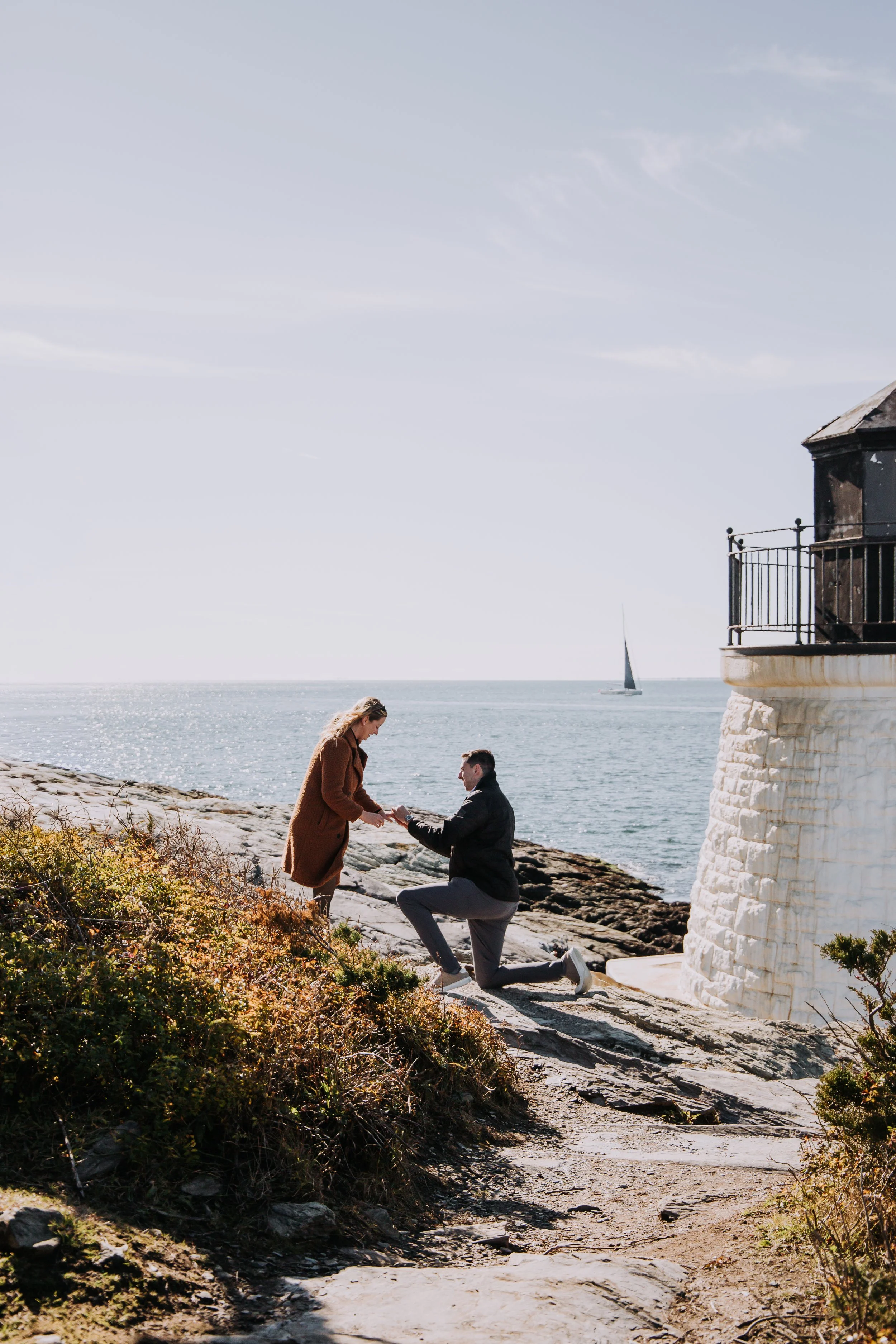 A man proposing marriage to a woman on a rocky shoreline near the ocean, with a sailboat in the background and a lighthouse on the right.