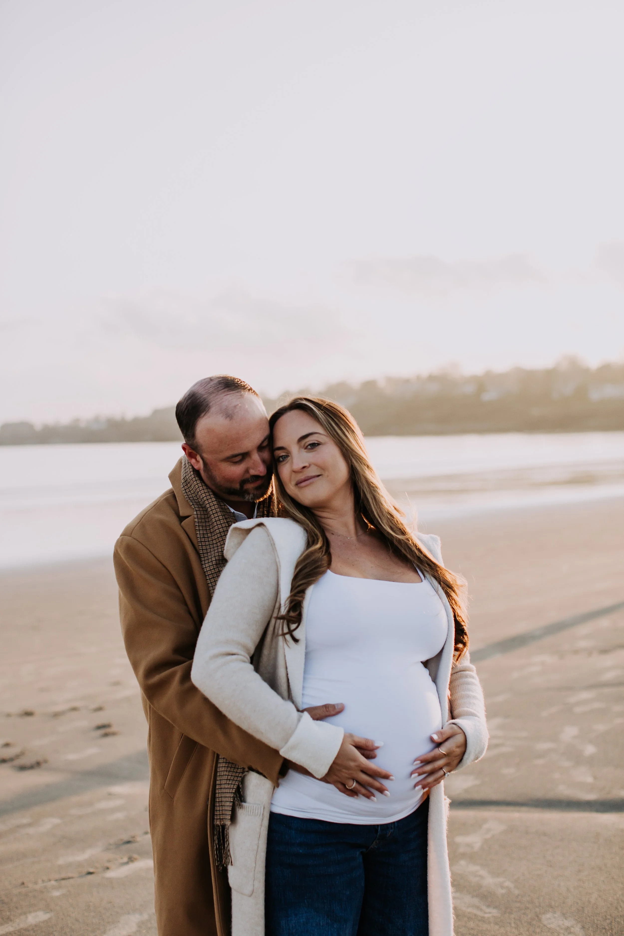A pregnant woman and a man standing on a beach at sunset, with the man hugging the woman from behind and both smiling.
