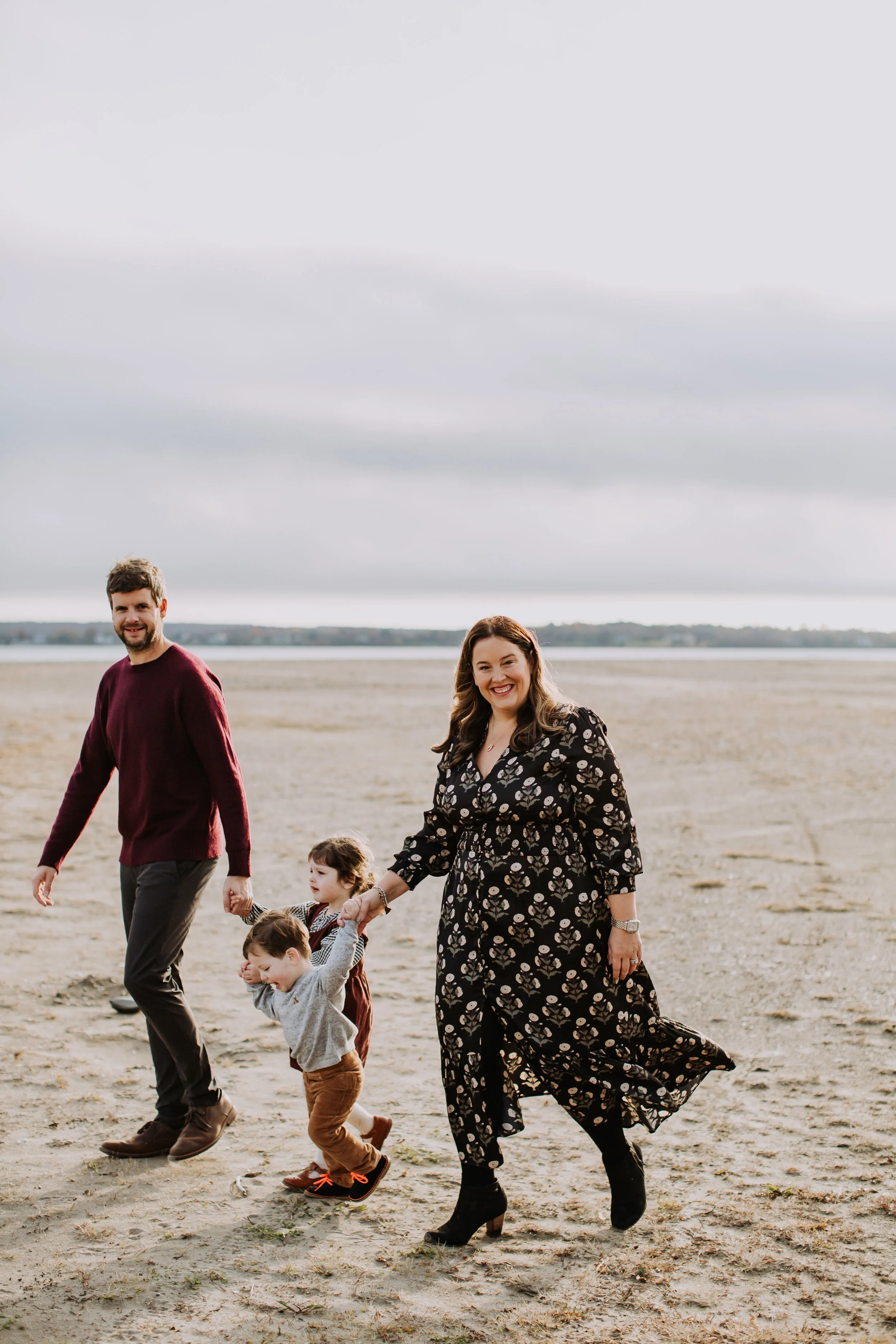 A happy family of four walking on a sandy beach, smiling, holding hands, with a cloudy sky in the background.