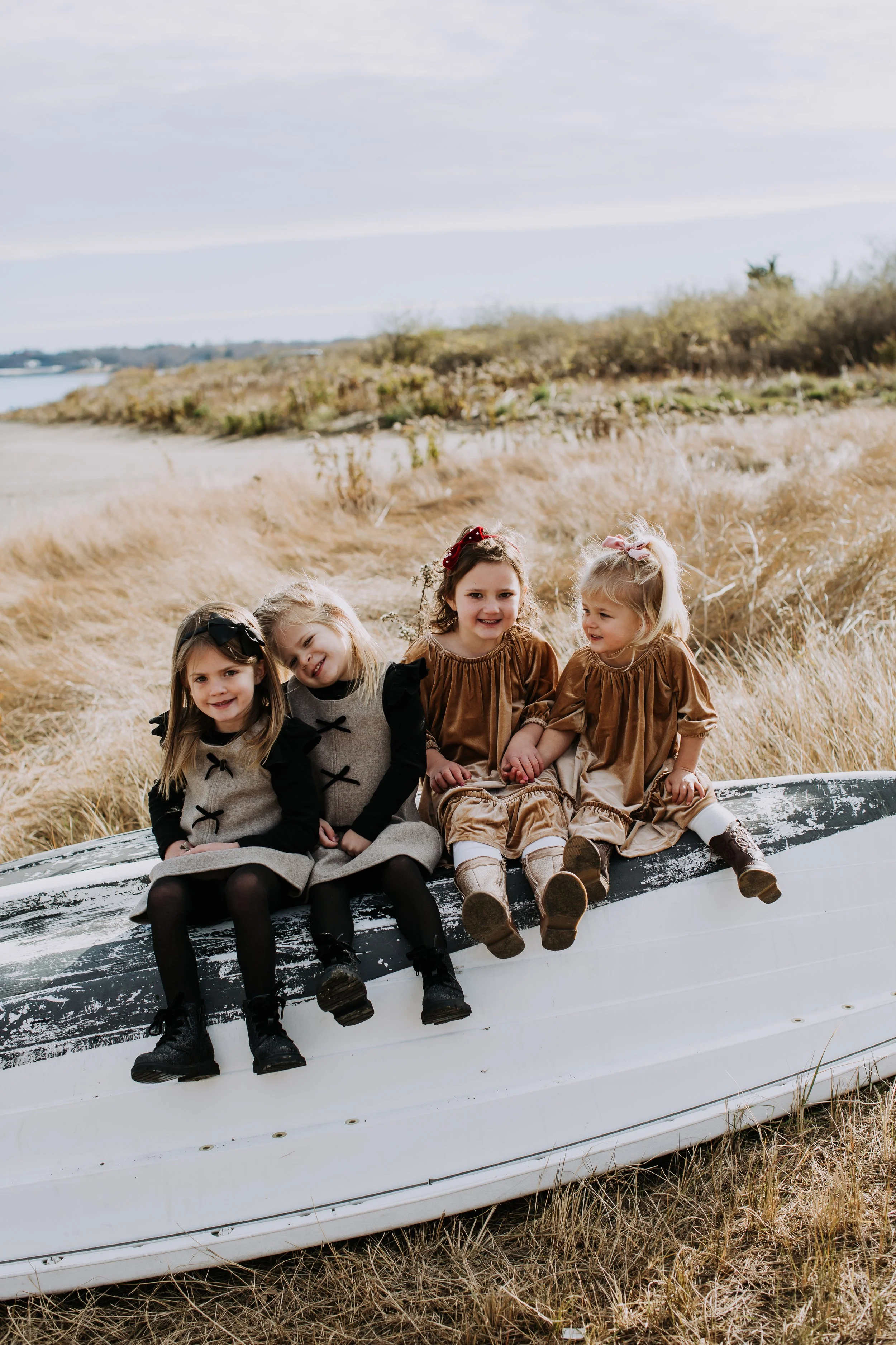 Four young girls sit on an old rowboat in a field with tall grass, smiling and holding hands, with a body of water and cloudy sky in the background.