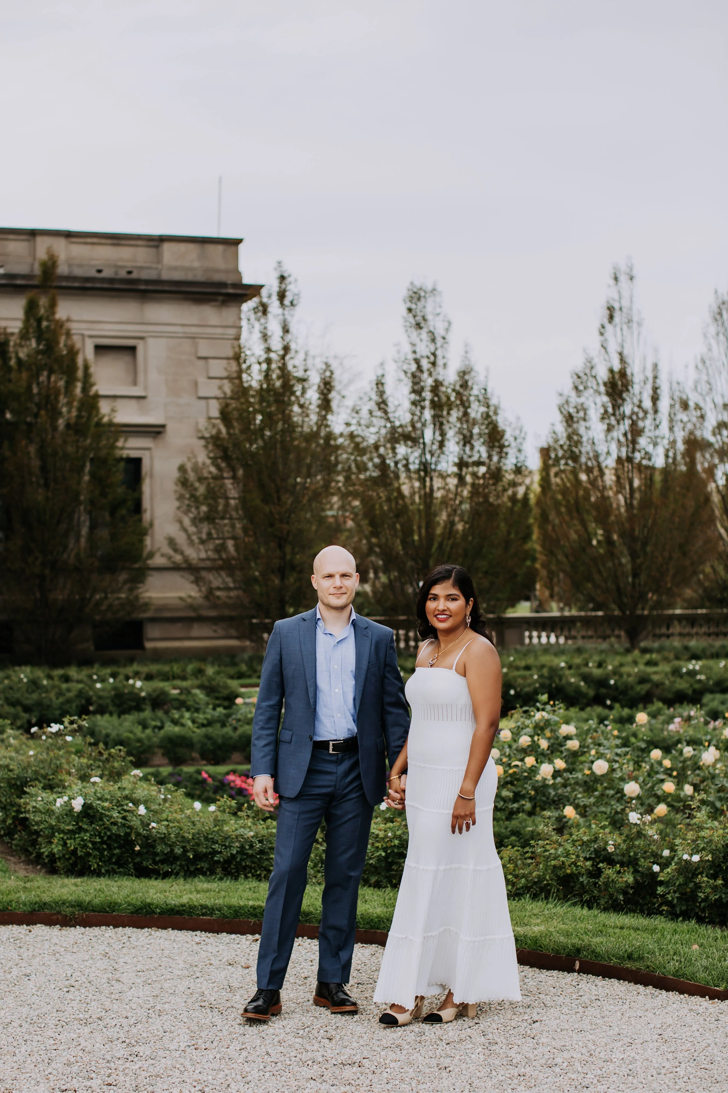 A couple dressed formally standing outdoors in front of a garden of white and pink roses with trees and a historic building in the background.