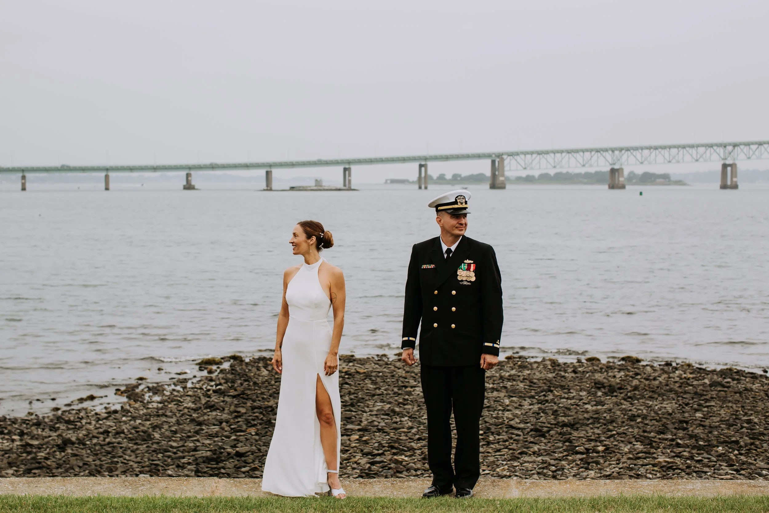 A bride in a white sleeveless wedding dress with a high slit and a groom in a navy naval officer uniform, standing on a grassy area by the water with a bridge in the background.