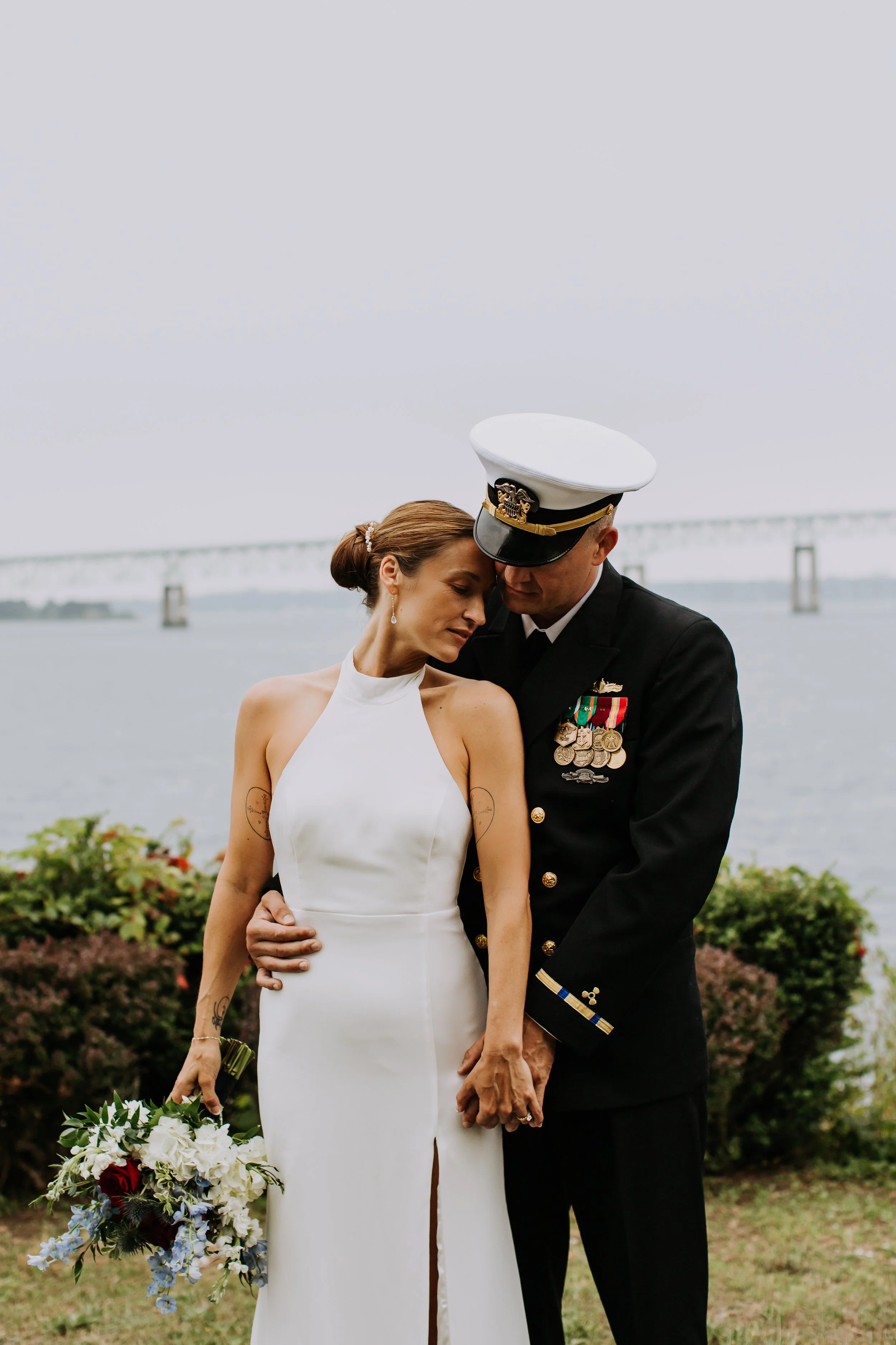 A couple dressed in wedding attire, with a woman in a white wedding gown holding a bouquet and a man in a navy military uniform with medals, embracing outdoors near a body of water with a bridge in the background.