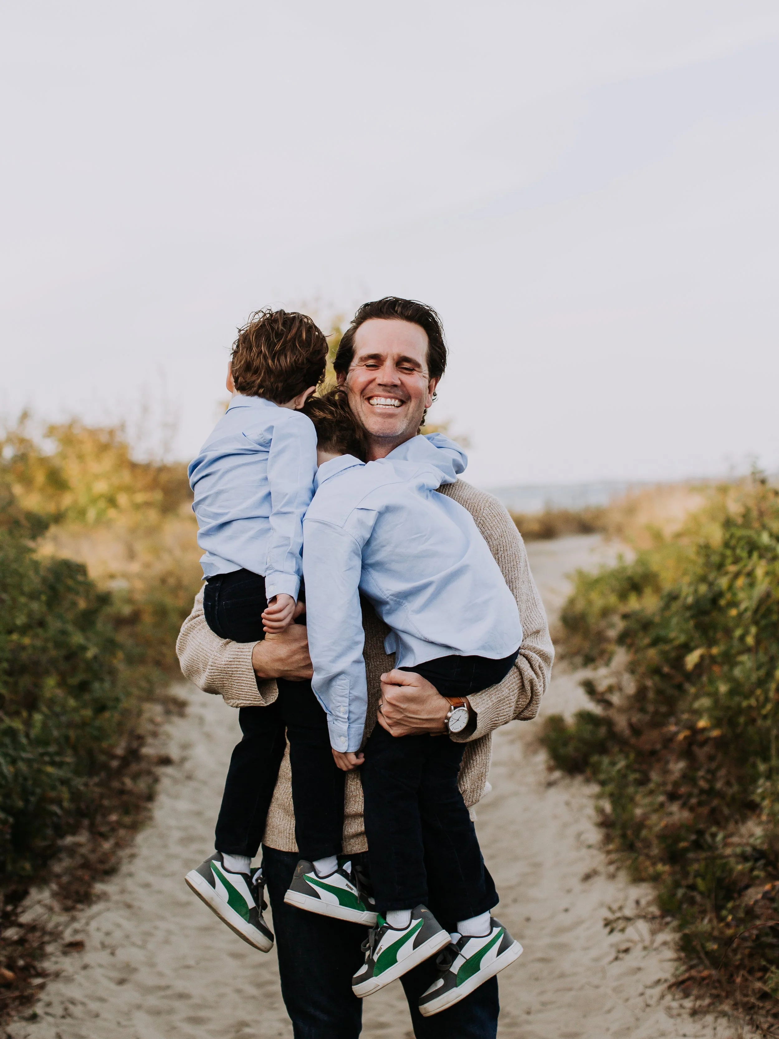 A man holding two children, all dressed in light blue shirts, on a sandy path with greenery on either side, smiling happily.
