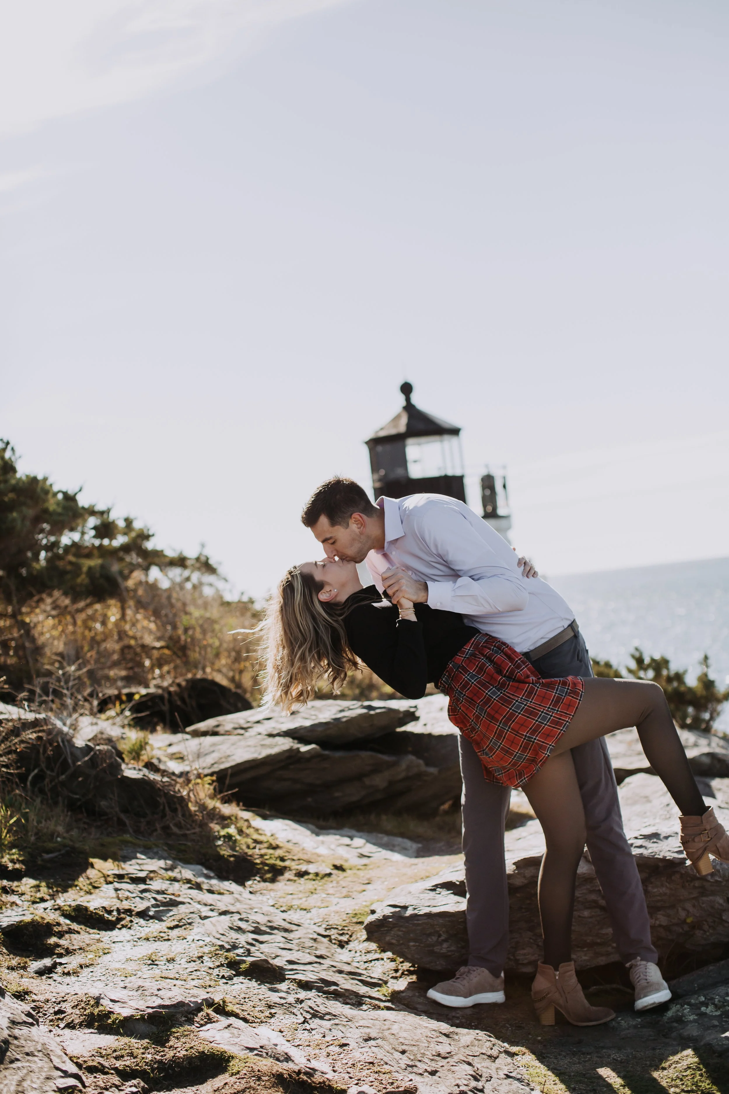 A couple kissing on a rocky beach with a lighthouse in the background