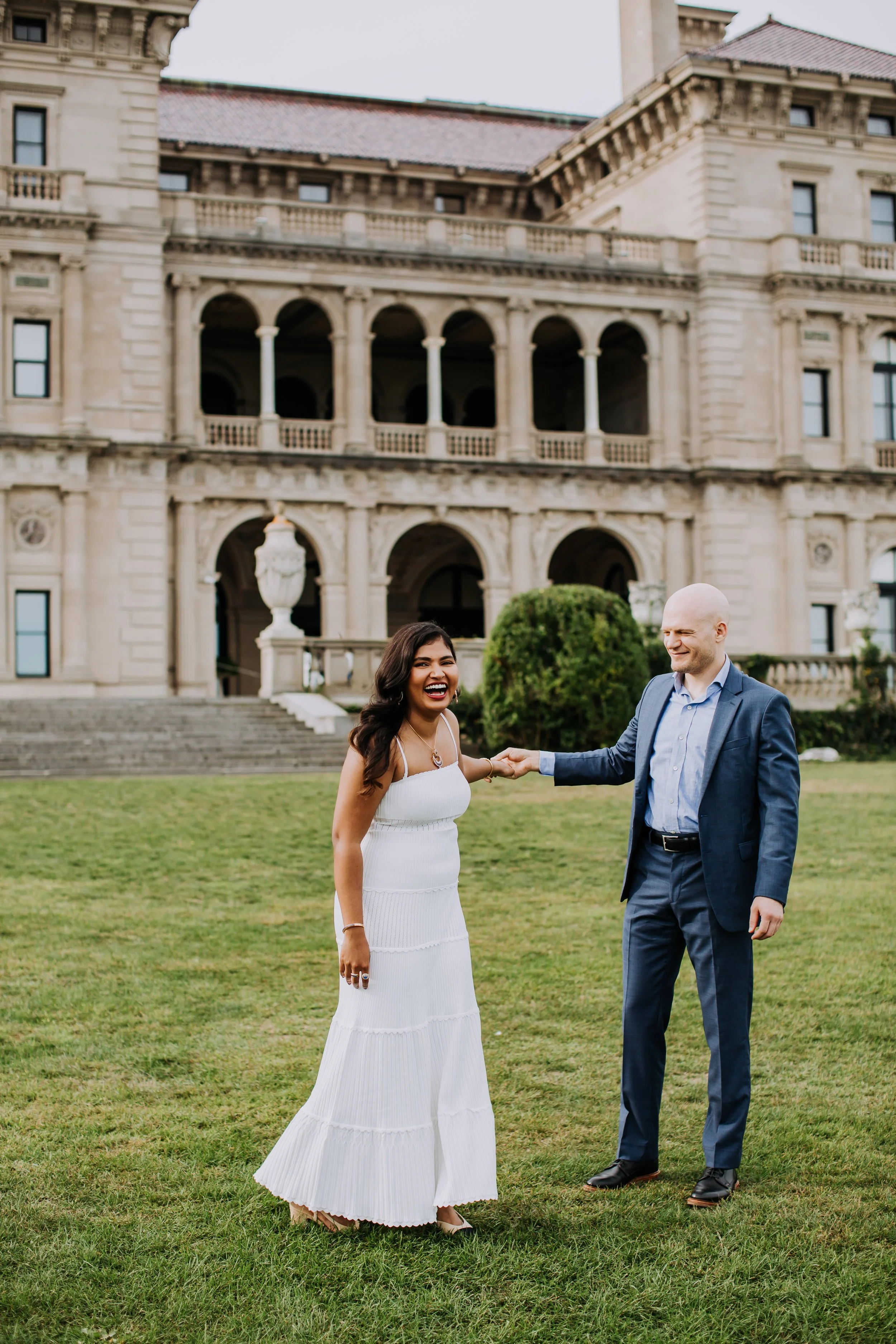 A woman and a man standing on grass in front of a large, historic stone mansion with columns and arched windows. The woman is wearing a white dress and laughing, while the man, dressed in a blue suit, is holding her hand and smiling.
