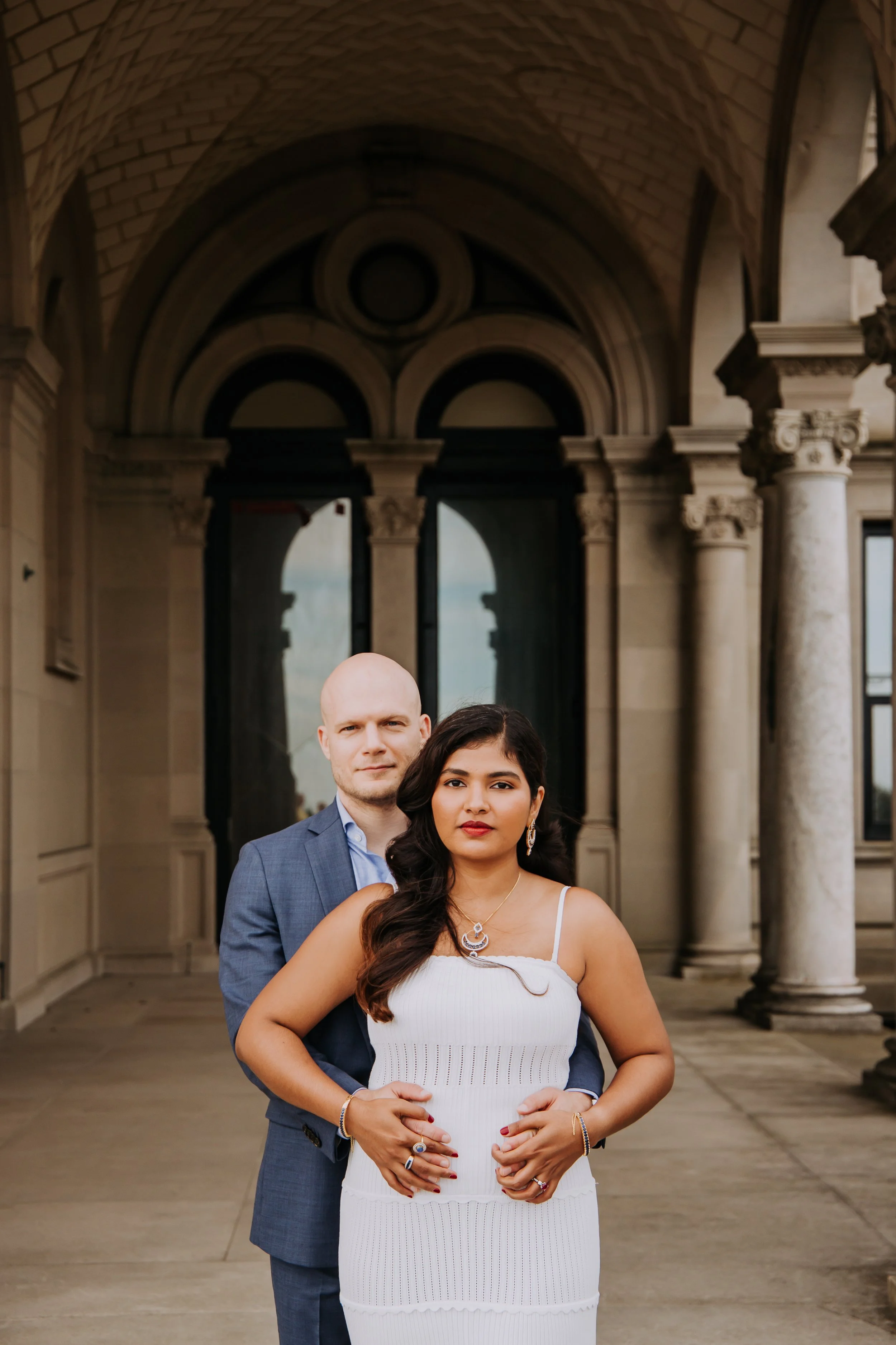 A couple standing in front of a grand historic building with columns and arches. The woman is wearing a white dress and jewelry, and the man is dressed in a blue suit.