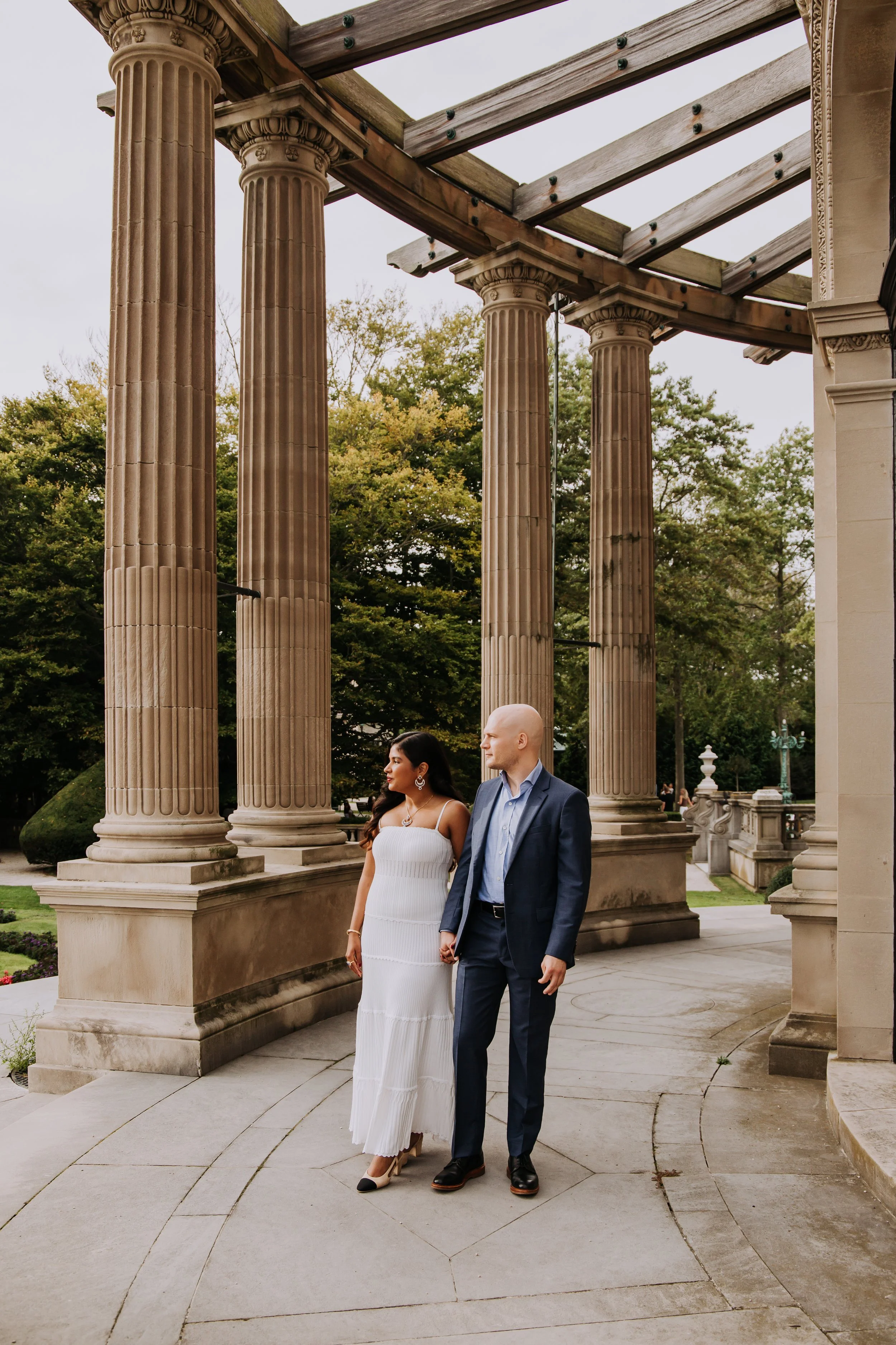 A couple holding hands, standing under a historic building with large classical columns, surrounded by trees and an overcast sky.