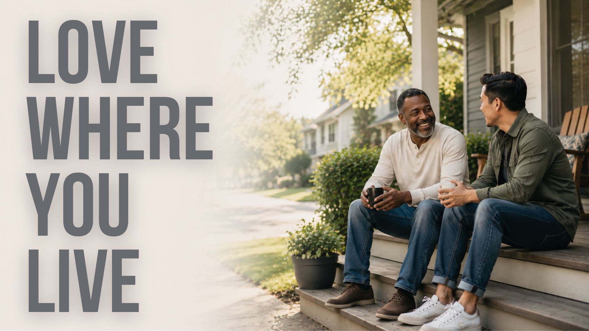 Two men sitting on a porch talking and smiling, each holding a coffee mug, with a suburban neighborhood in the background and a large tree overhead.