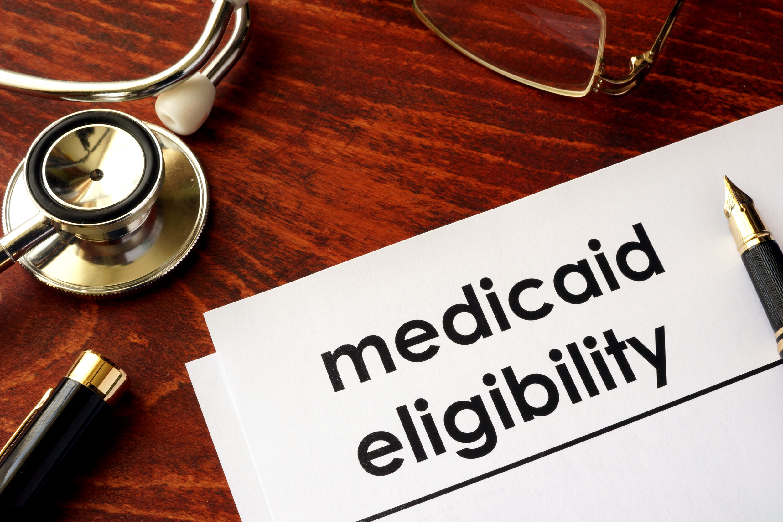 Estetoscopio y gafas sobre una mesa junto a un papel y un bolígrafo que dice "Medicaid Eligibility"