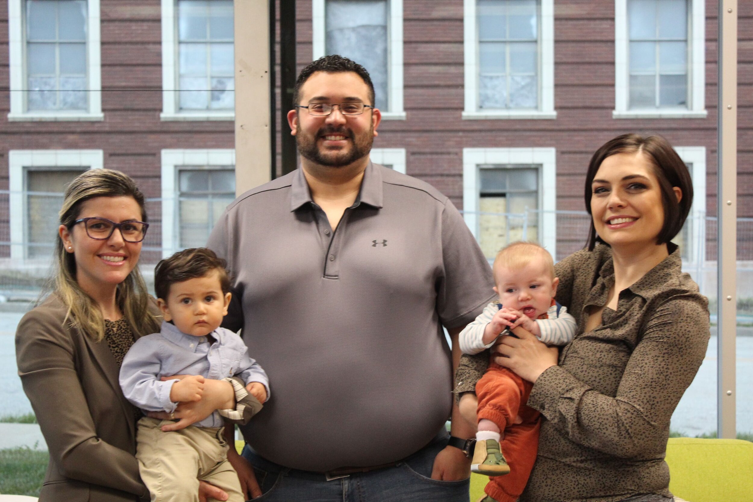 El personal de la KAN de pie frente a un gran ventanal con sus hijos en brazos. De izquierda a derecha: Carolina Biagi con su hijo en brazos, Ricardo Ortiz en el centro y Molly Gotobed con su hijo en el extremo derecho.
