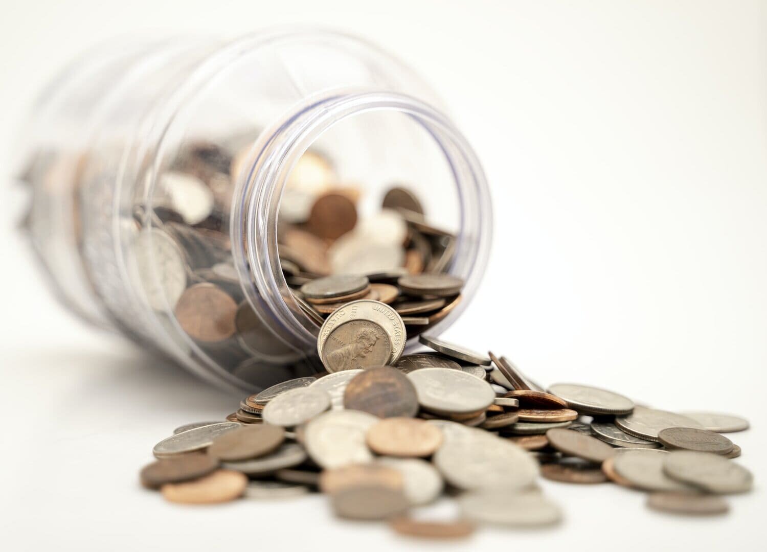 Un tarro de cristal lleno de monedas americanas tumbado de lado, derramando su contenido sobre una superficie blanca que sirve de metáfora del gasto de dinero.